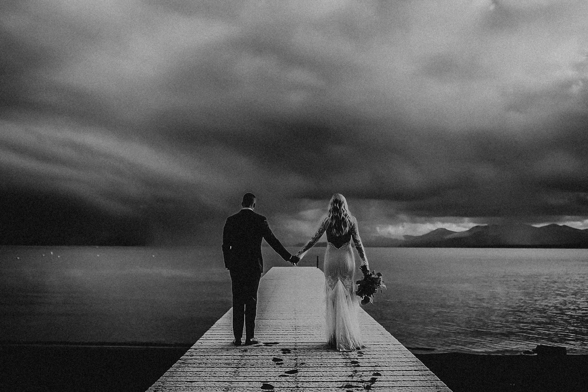 A black-and-white photo of a bride and groom walking hand in hand on a wooden pier by the water, with dark, cloudy skies and mountains in the distance.