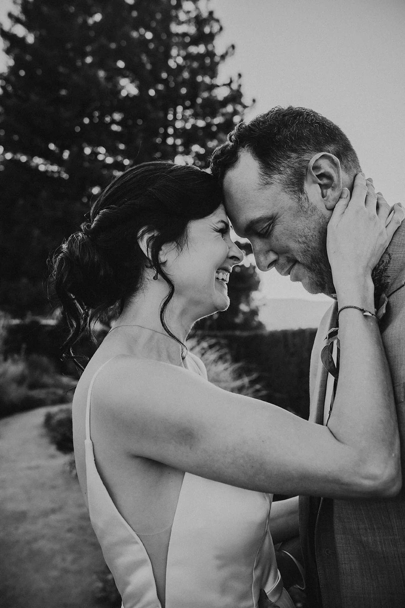 A black and white photo of a couple with foreheads touching, smiling and embracing outdoors.