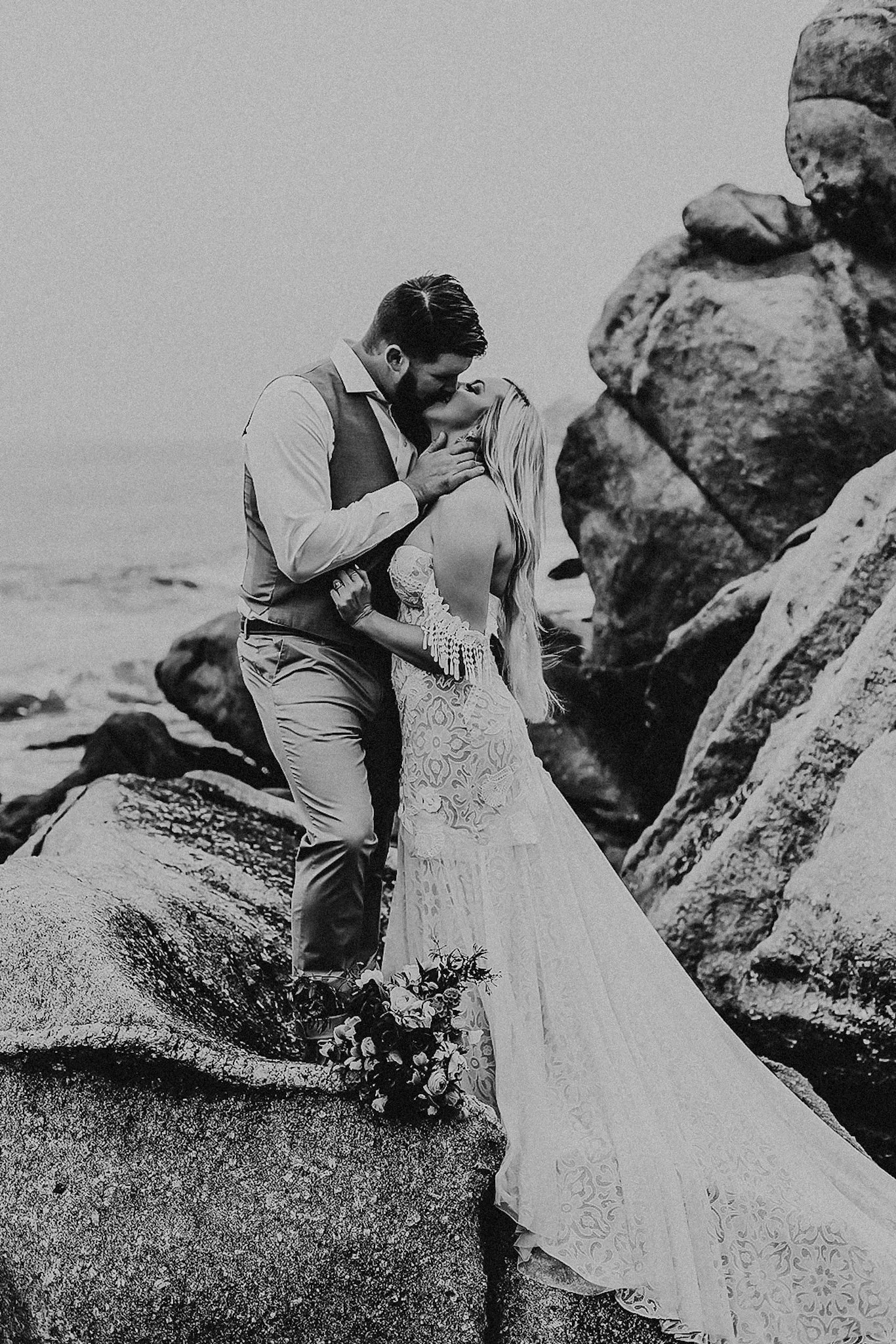 A black and white photo of a couple in wedding attire sharing a kiss on rocks by the ocean.