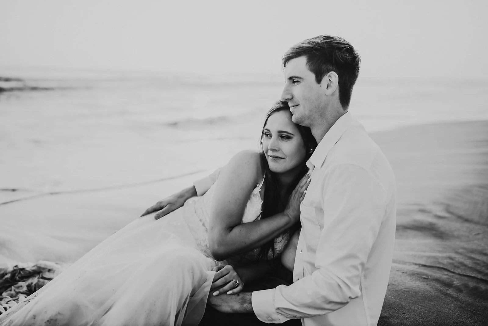 A black-and-white photo of a couple sitting on the beach, with the woman resting her head on the man's shoulder and looking into the distance, the man looking forward, both appearing tender and contemplative.