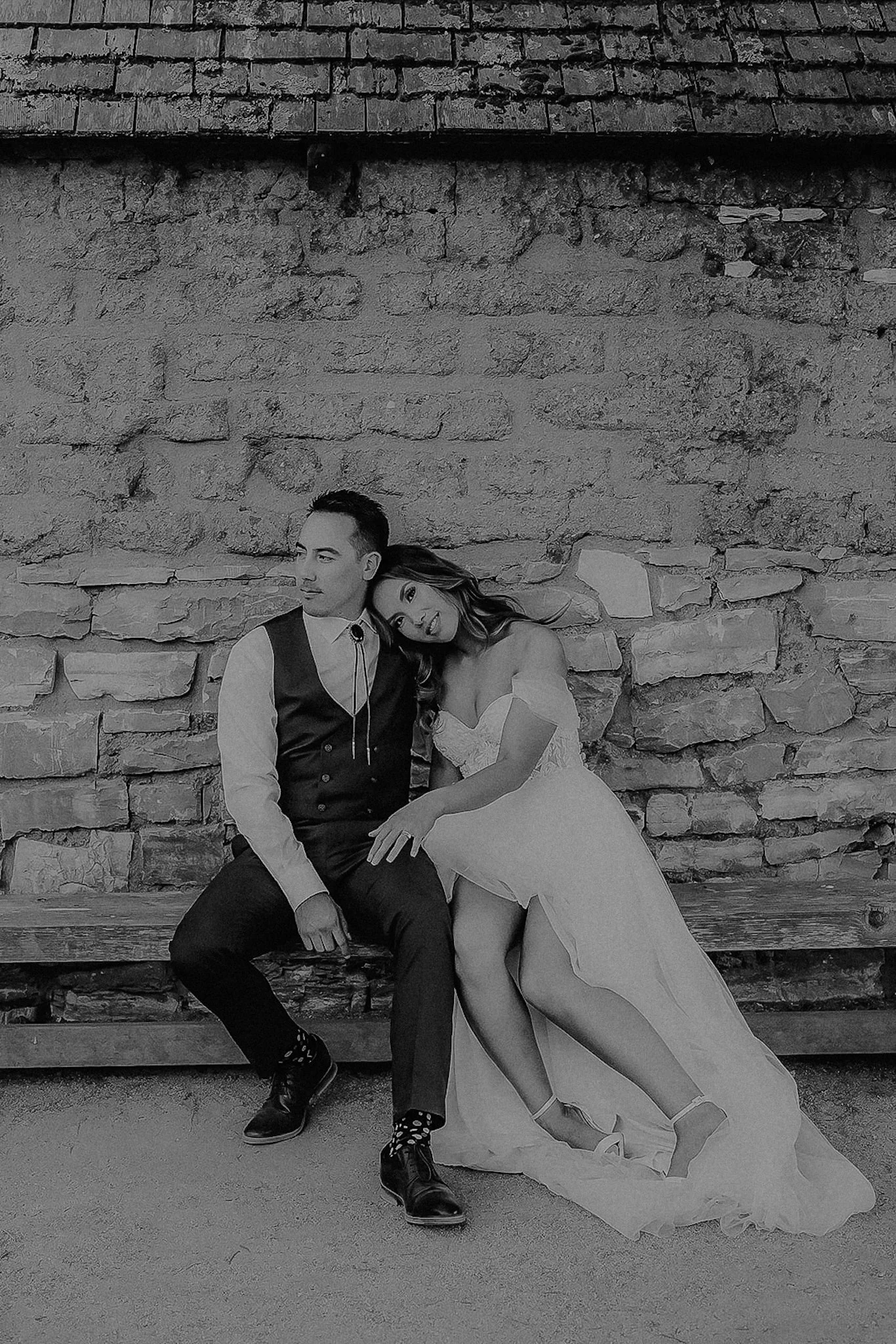 A black-and-white photo of a couple sitting on a wooden bench against a stone wall. The woman is leaning her head on the man's shoulder, wearing a wedding dress with an off-the-shoulder style, and the man is dressed in a vest, shirt, and tie.
