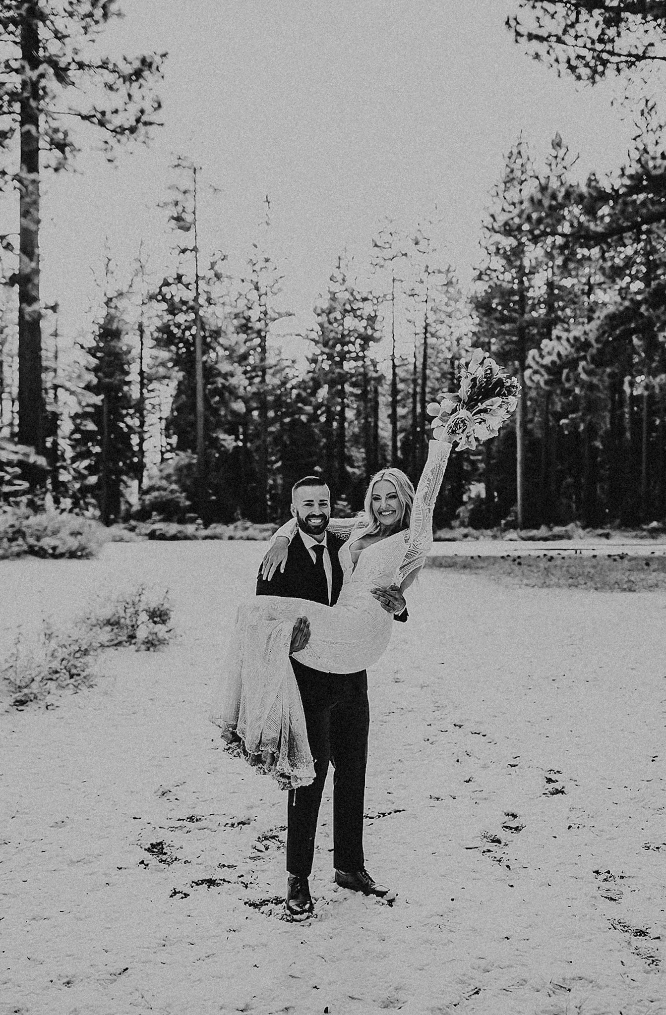 A black and white photo of a happy couple outdoors in the snow, with the man carrying the woman who is holding a bouquet with one arm raised.
