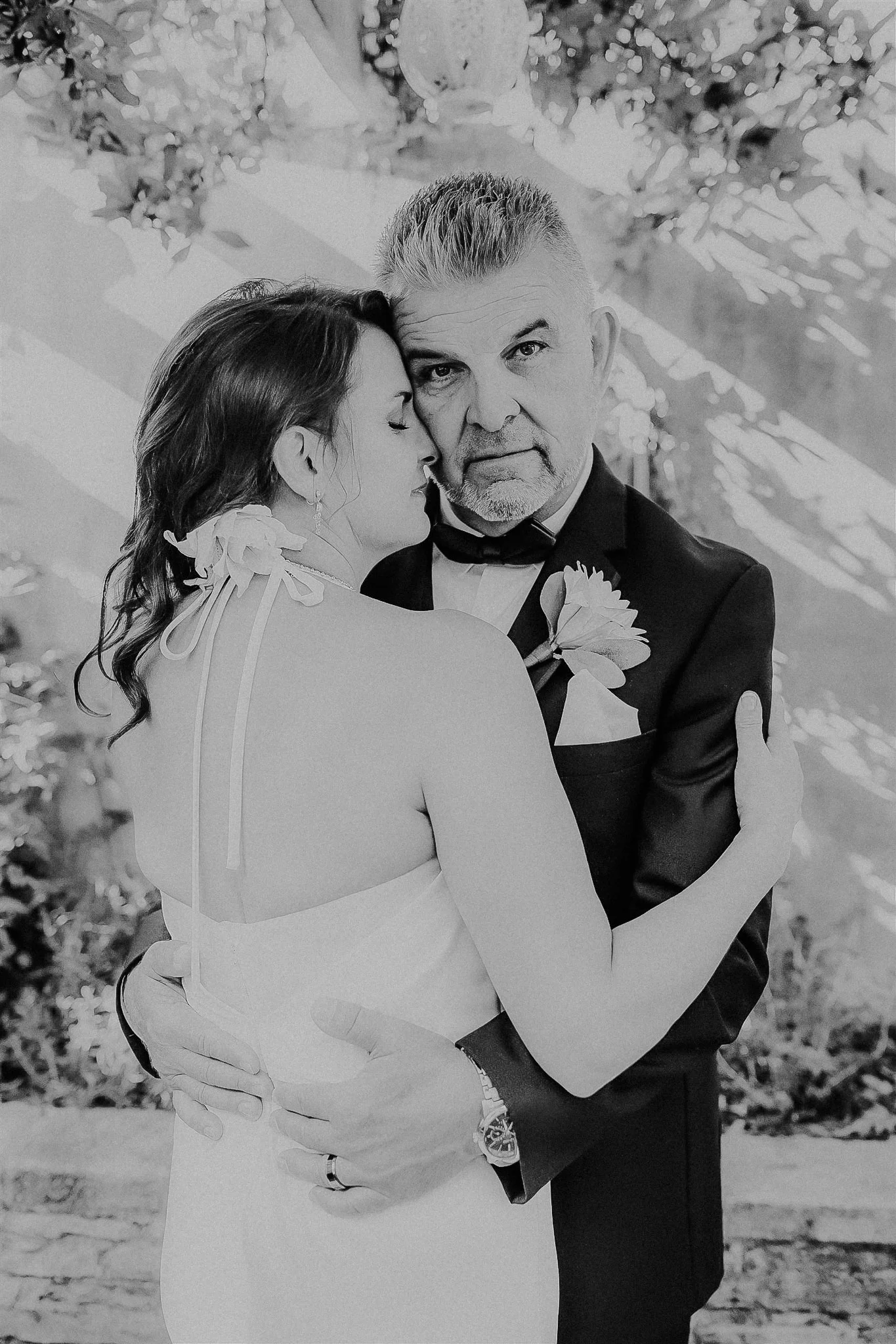 A black-and-white photo of a bride and an older man, possibly her father, embracing each other. The bride is wearing a sleeveless dress with a bow detail on her shoulder, and the man is wearing a tuxedo with a boutonniere. They are outdoors with blur