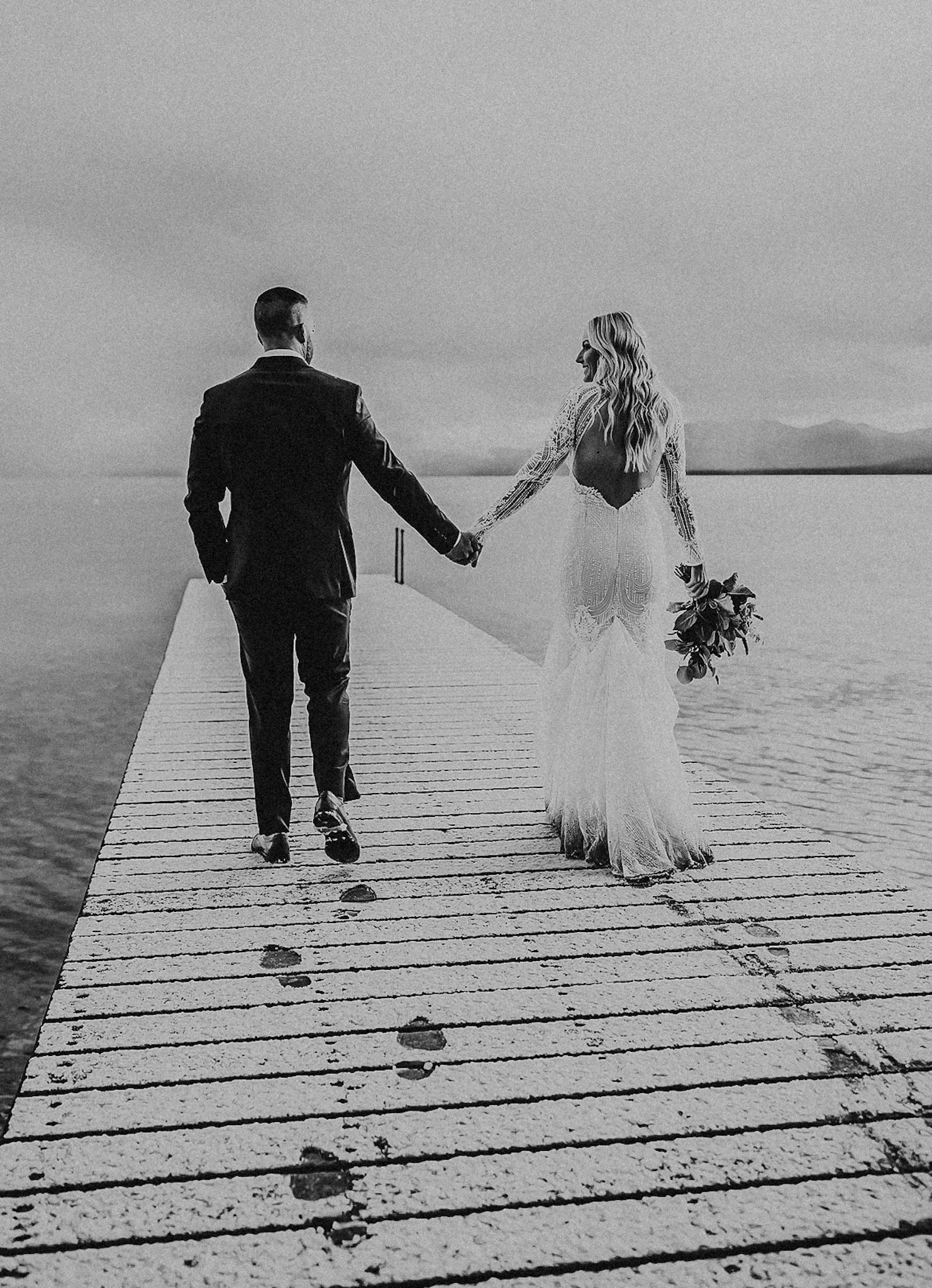 Black and white photo of a couple walking hand in hand on a dock over water, with mountains in the background. The woman is wearing a long wedding dress and holding a bouquet, and the man is dressed in a suit.