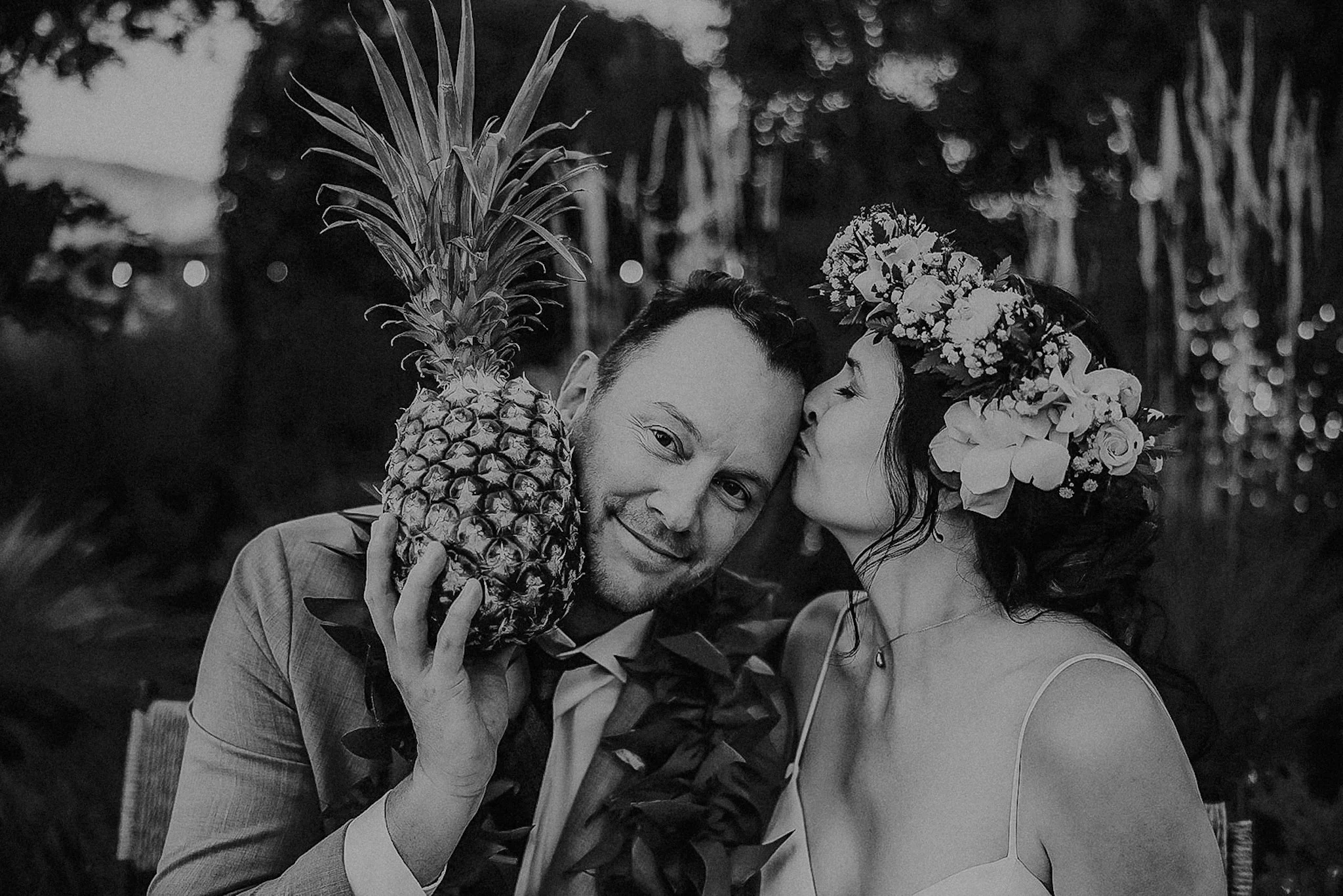 A woman wearing a flower crown kisses a man on the cheek while holding a pineapple, both smiling, outdoors with trees in the background.