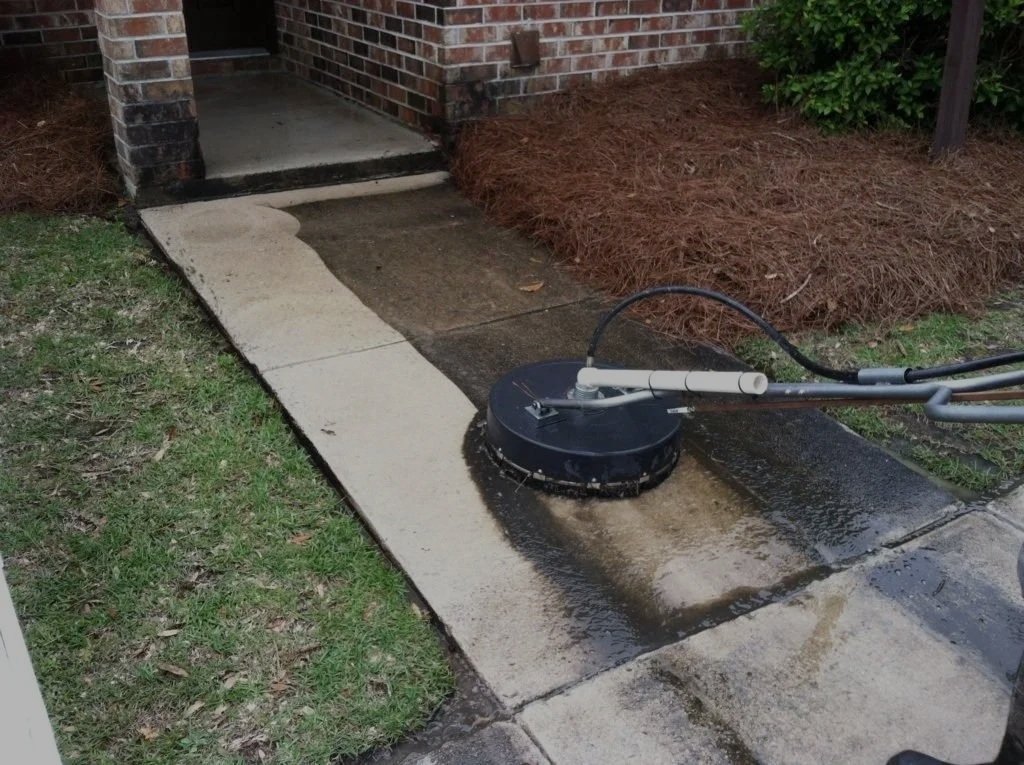 Power washer cleaning a concrete sidewalk and porch area outside a brick house.