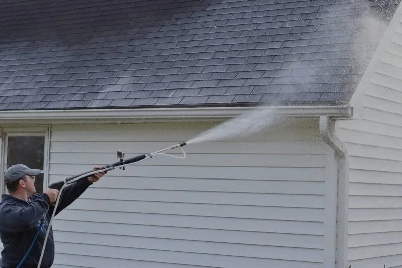 A person pressure washing the exterior wall of a house with a hose and a spray wand.