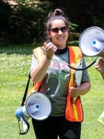 Young woman holding two megaphones outdoors on a sunny day, wearing sunglasses and a safety vest.