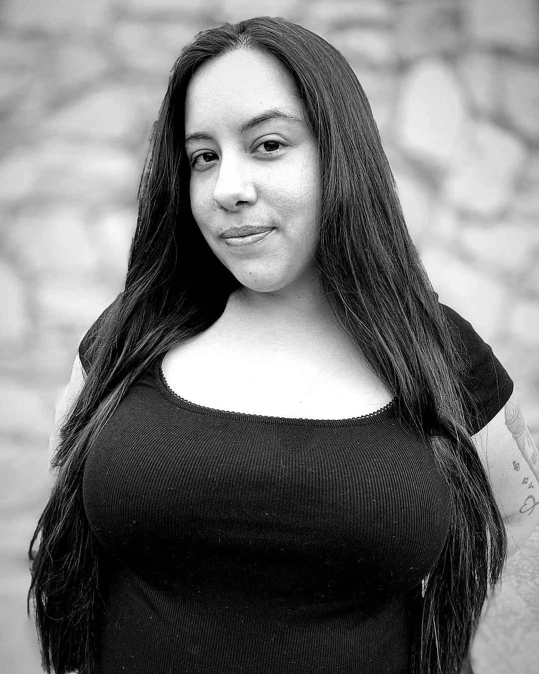 Black and white portrait of a woman with long dark hair, wearing a black top, standing outdoors with a blurry brick wall background.