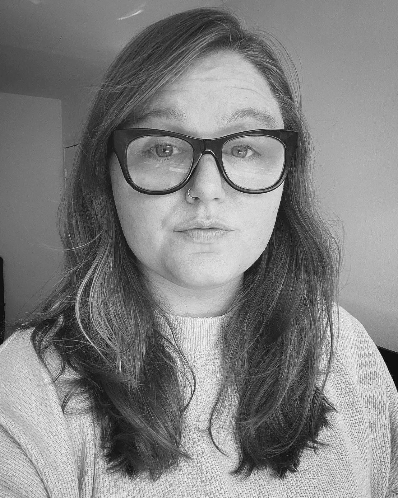Black and white portrait of a person with long wavy hair wearing large glasses and a nose ring, looking directly at the camera.