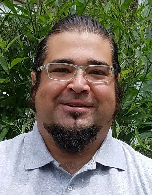 Close-up of a man with long dark hair, glasses, and facial hair, smiling, standing in front of green foliage.