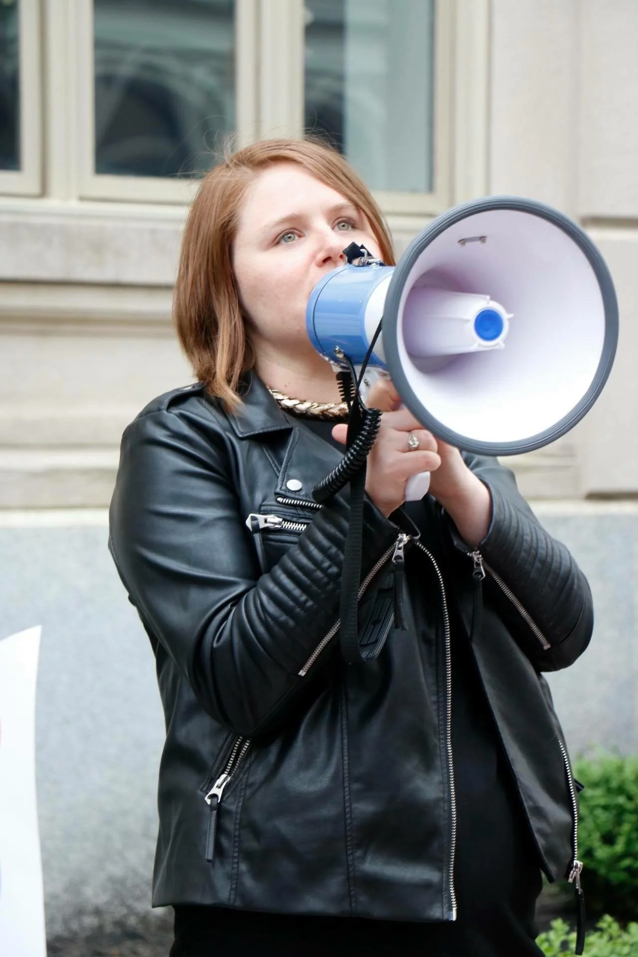 Person with red hair holding a blue and white megaphone, wearing a black leather jacket, speaking at a rally or protest outside near a building.