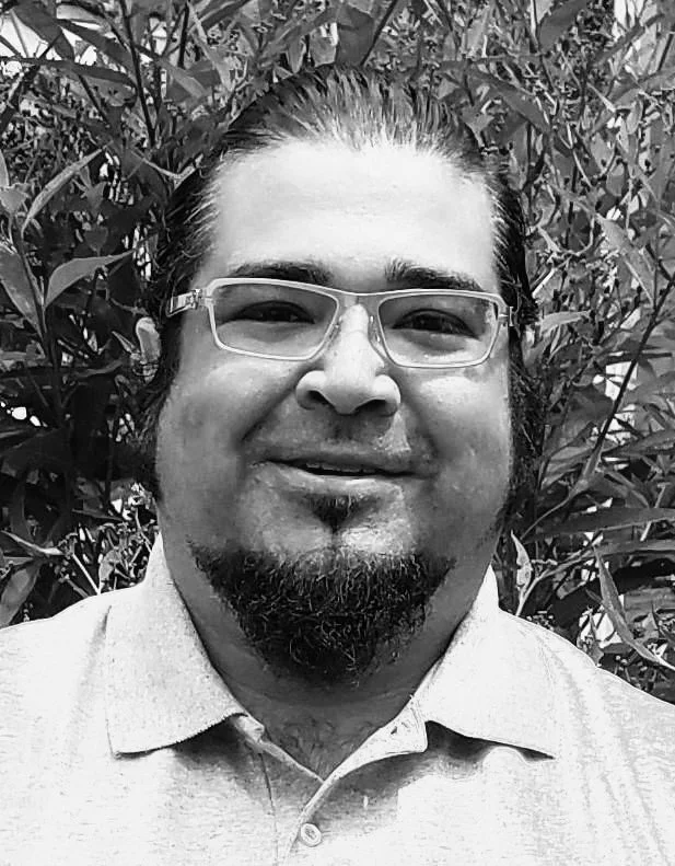 A man with glasses, a goatee, and a beard, smiling, standing outdoors in front of foliage.