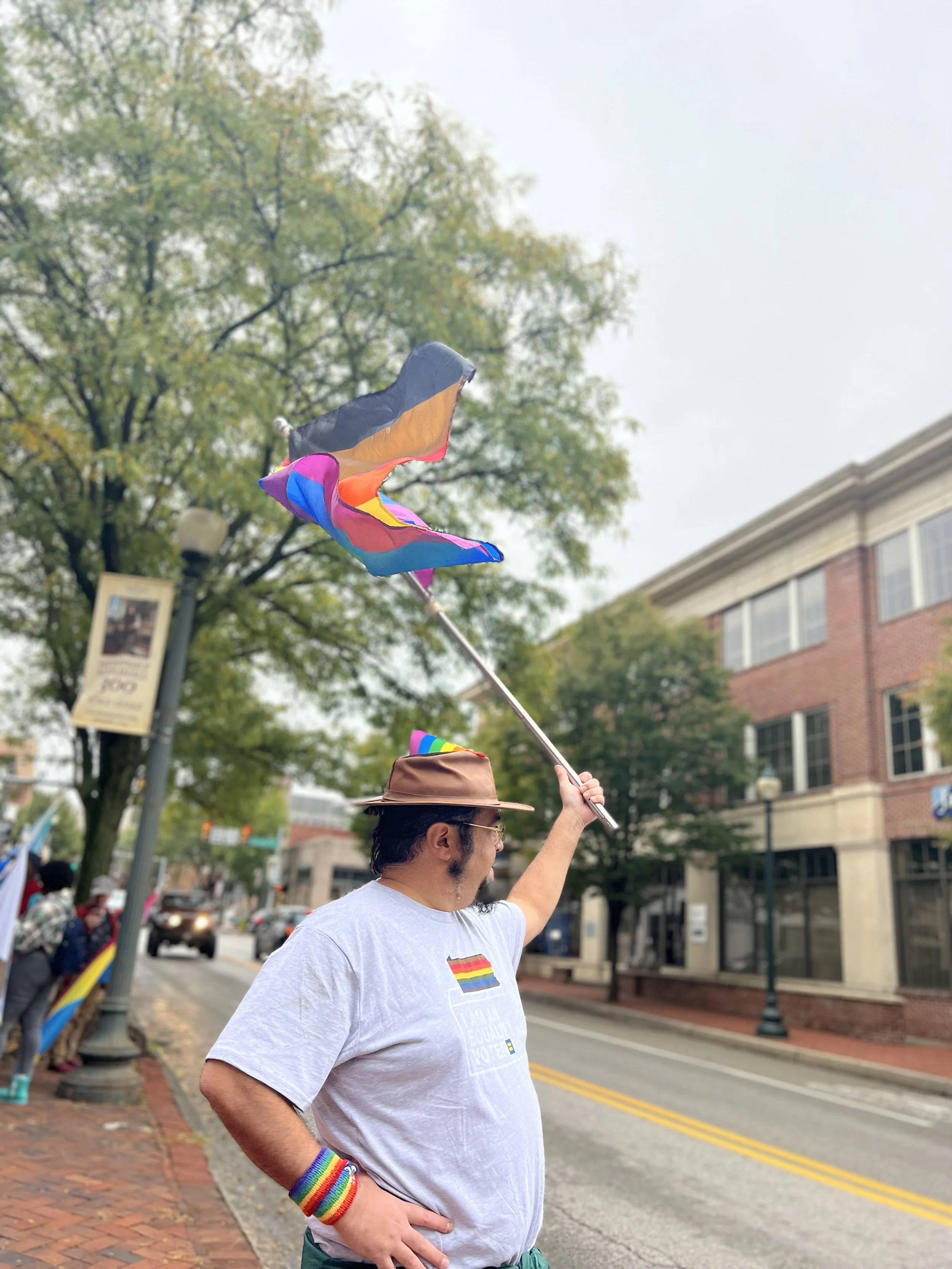 A man wearing a white T-shirt with a rainbow pride flag design and rainbow wristbands waving a rainbow pride flag at a pride parade on a city street.