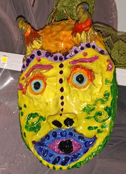 Colorful, artistic mask with bright yellow face, large blue eyes, purple eyebrows, and an exaggerated mouth with pink and purple lips. Decorative elements include green swirls, purple dots, and orange and red accents on top resembling hair or horns.