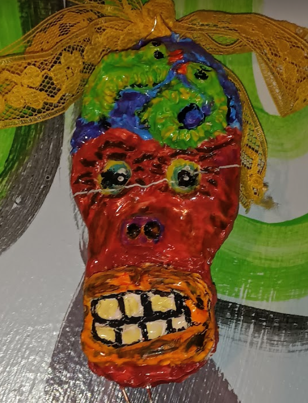 Colorful, hand-painted mask with exaggerated facial features, including large eyes, nose, and an open mouth with visible teeth, decorated with yellow lace and ribbons.