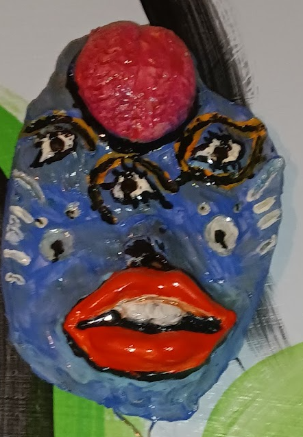 A painted mask with blue face, red lips, and three eyes, topped with a red strawberry.