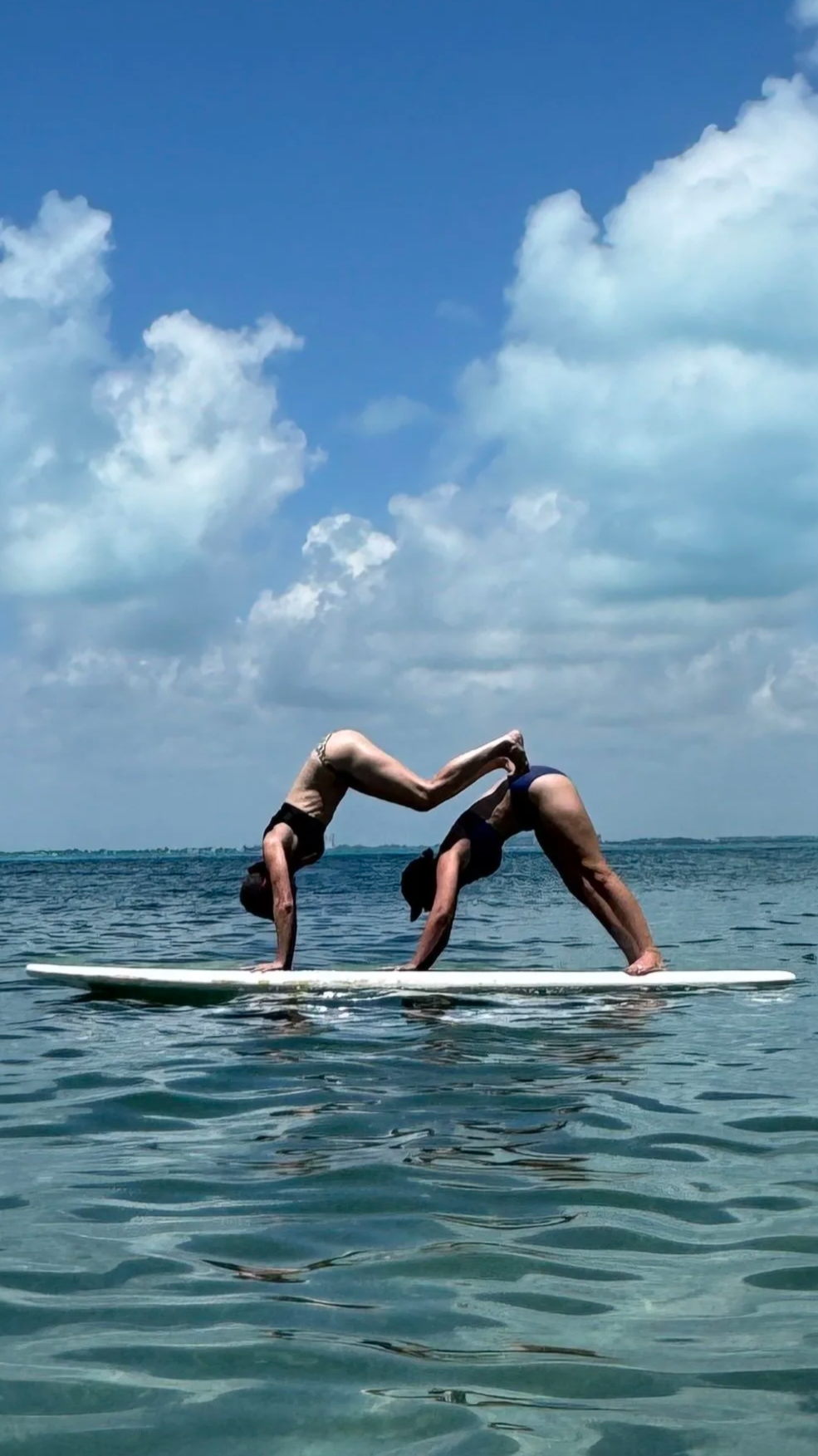 Two women practicing yoga on a paddleboard in the water under a partly cloudy sky.