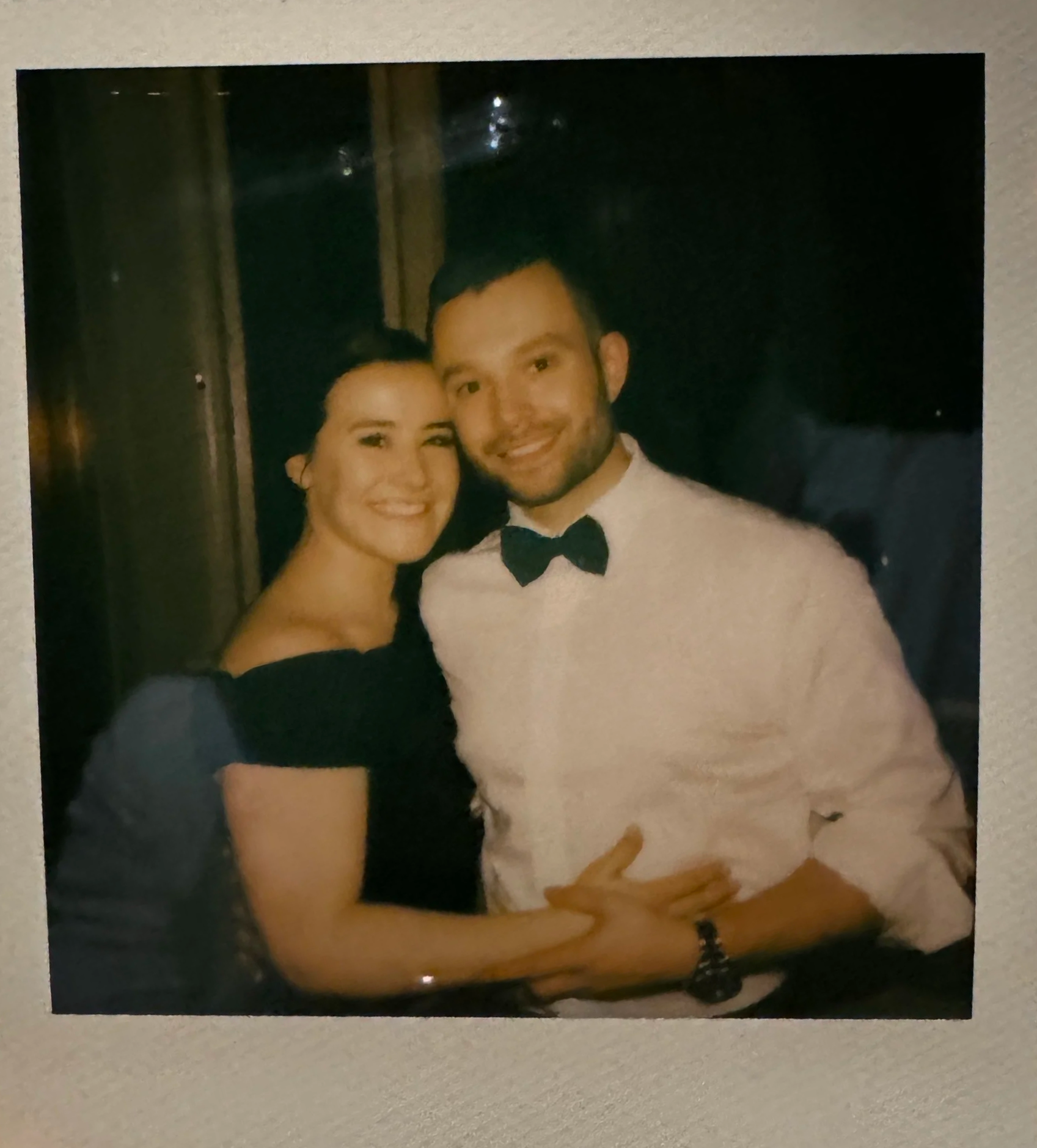 A smiling woman and man dressed in formal attire, with the woman in a black off-the-shoulder dress and the man in a white shirt with a black bow tie, posing together in a dark indoor setting.