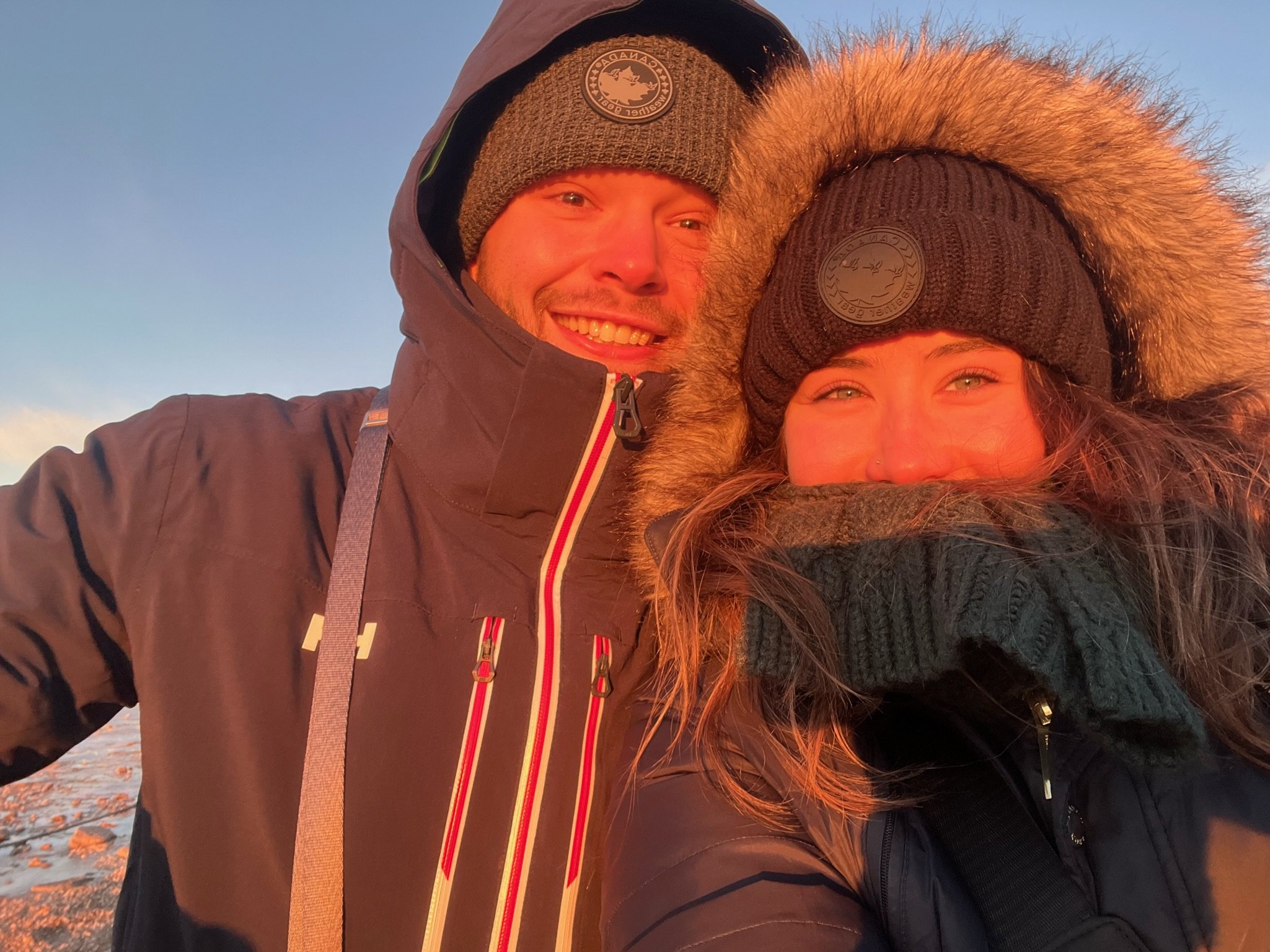 A man and woman outdoors in winter clothing, smiling at sunset, with snow-covered ground visible.