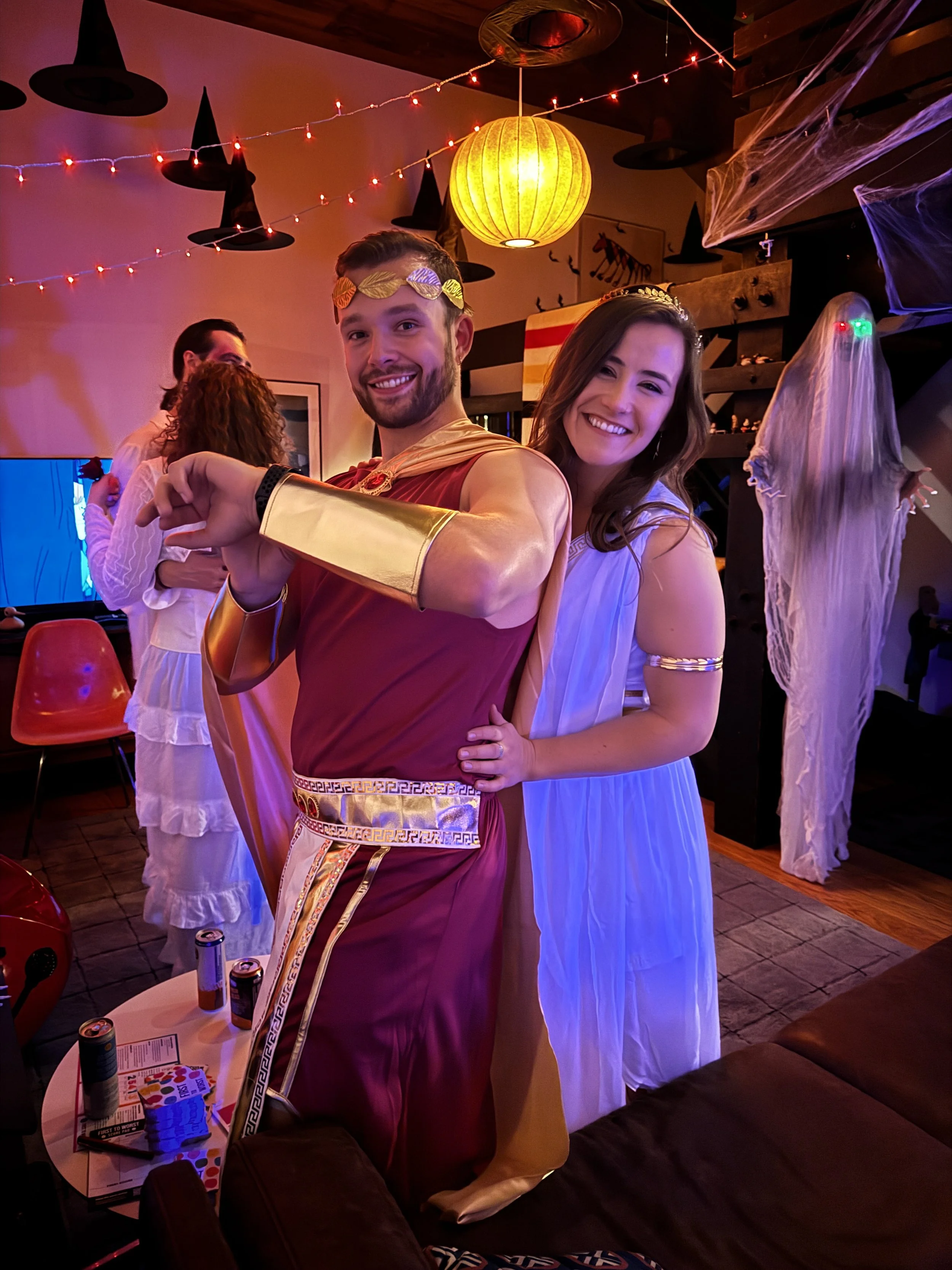 People dressed in Greek or Roman costumes at a Halloween party with decorations, string lights, and a ghost costume in the background.