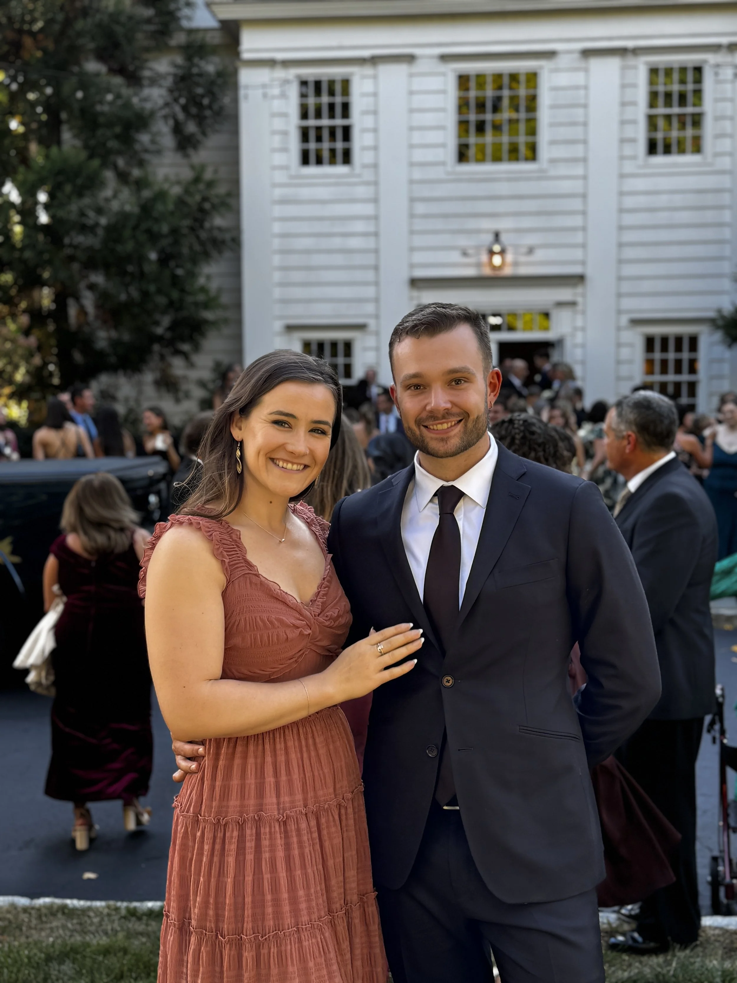 A smiling woman in a rust-colored dress and a man in a dark suit at an outdoor event in front of a white house with multiple windows and a crowd of people.
