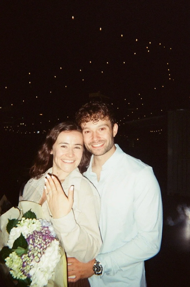 A young couple smiling and posing together at night. The woman has shoulder-length brown hair and is holding a bouquet of flowers. The man has curly dark hair and wears a white shirt and a watch. They are standing outdoors under a sky with star-like lights.