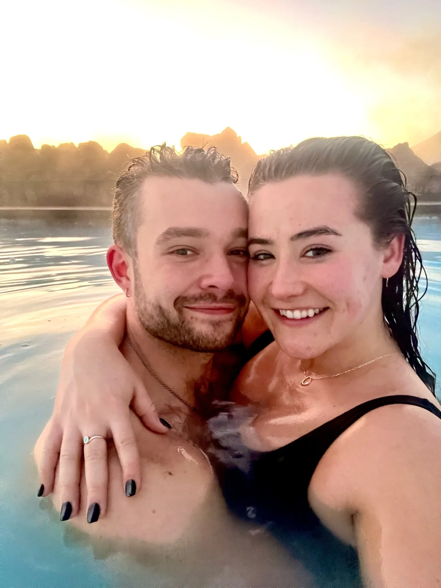 A smiling couple in a hot tub at sunset, with a scenic mountain view in the background.