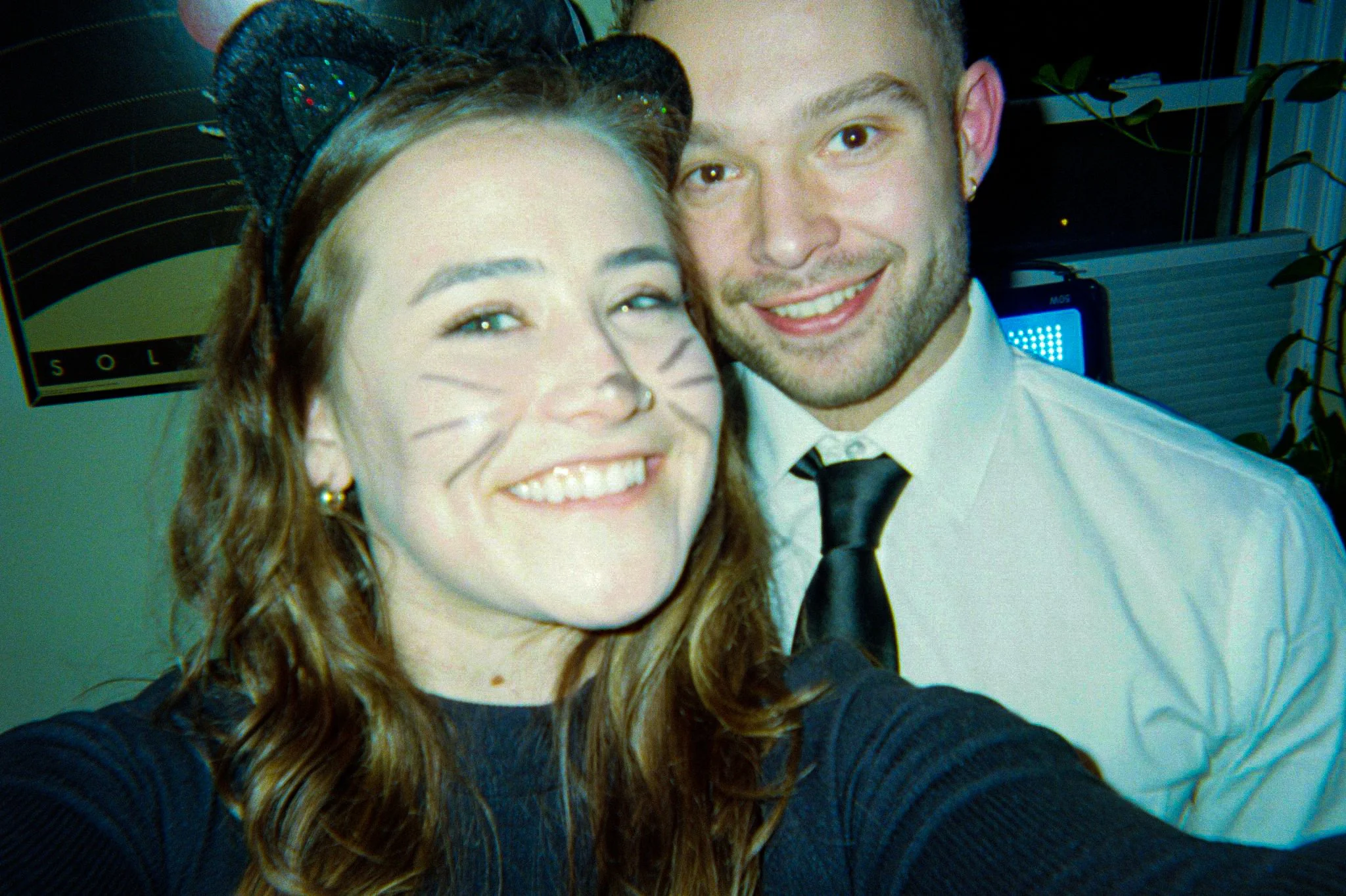 A young woman with makeup resembling a cat, wearing a headband with ears, and a nose and whiskers drawn on her face, taking a selfie with a young man in a white shirt and black tie.