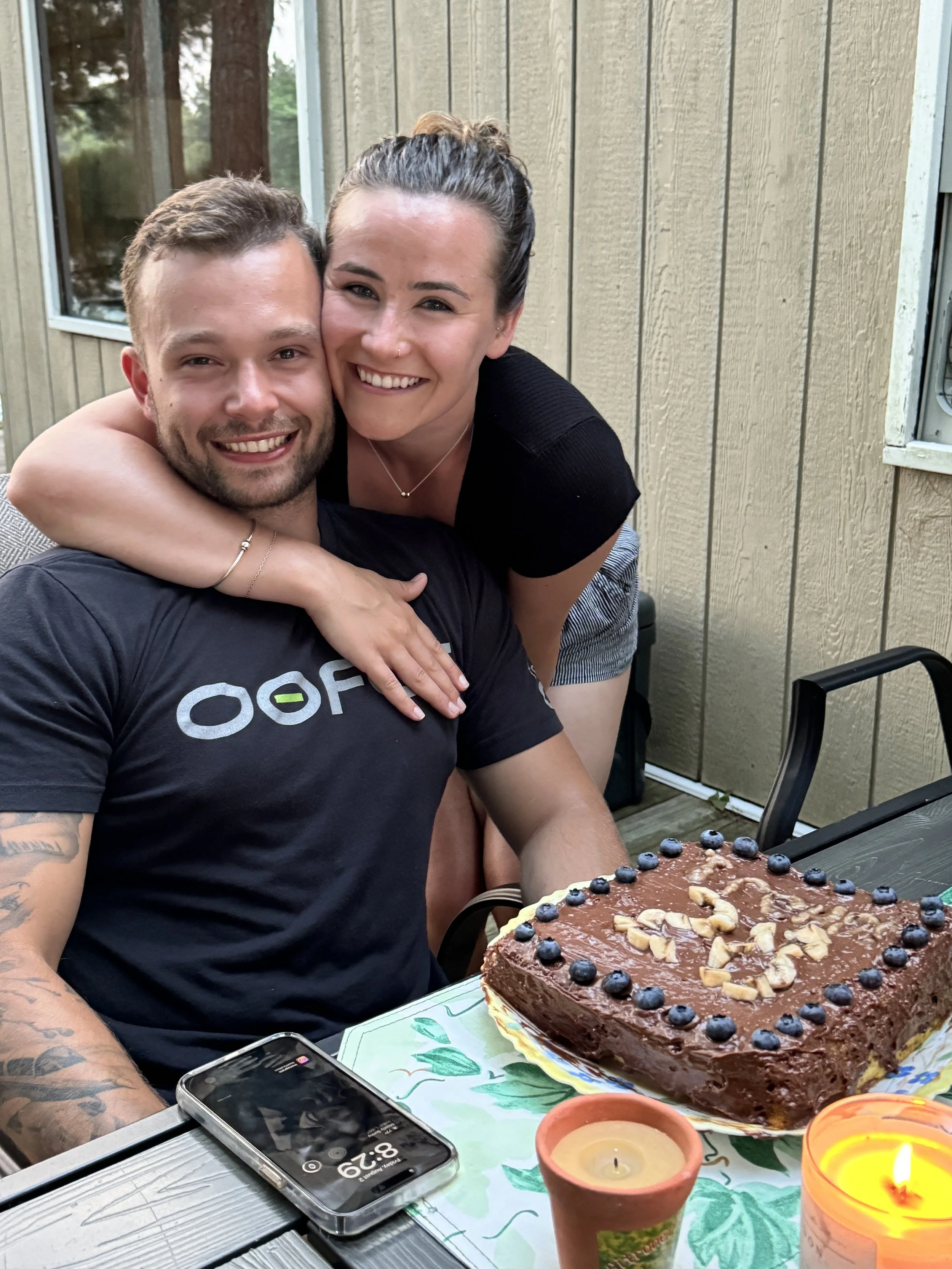 A woman and man smiling and hugging at a birthday celebration outdoors, with a chocolate cake with blueberries and nuts on the table, along with lit candles, a phone, and a floral tablecloth.
