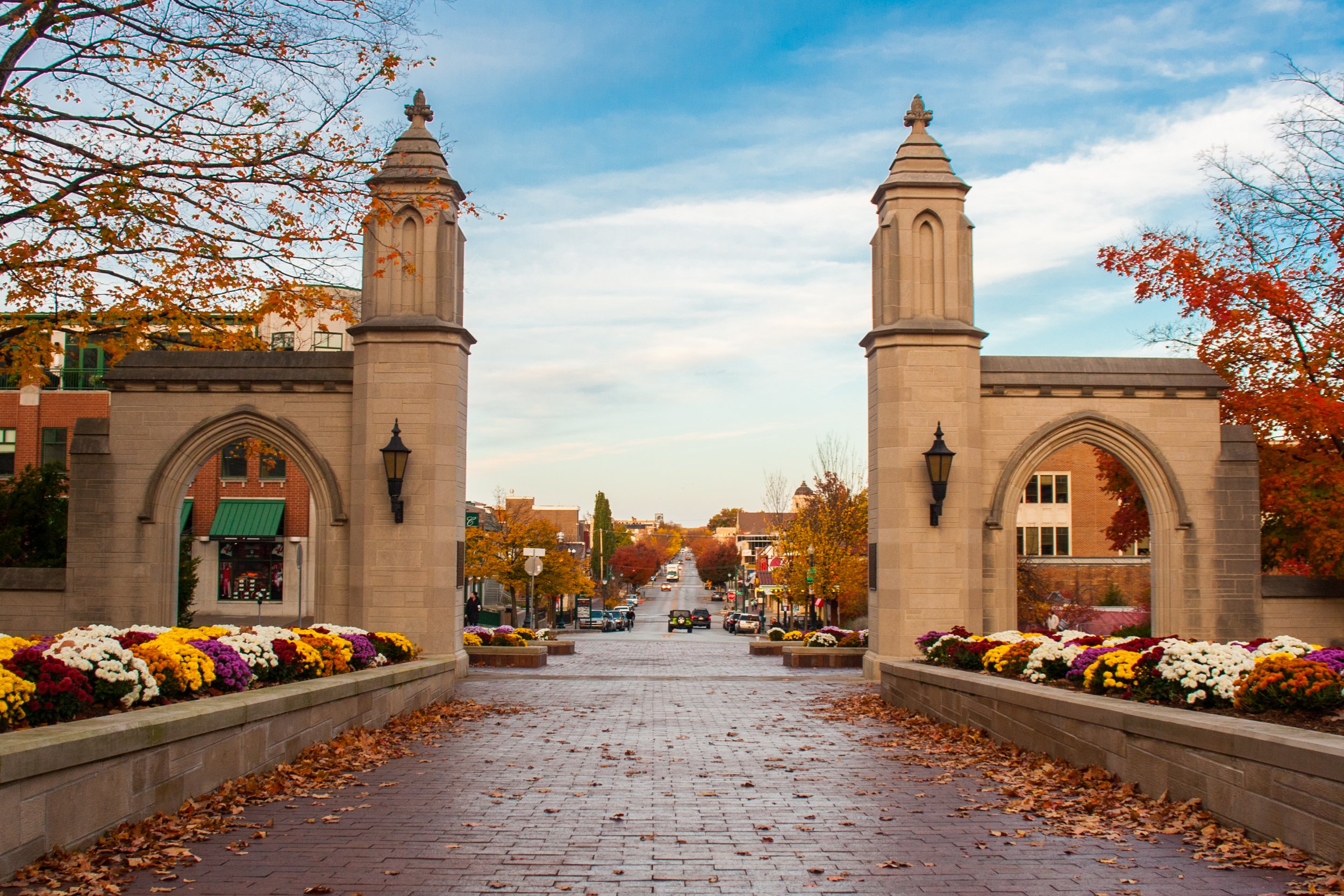 Stone archway entrance with two towers and flowerbeds with autumn-colored flowers, leading to a small town street with trees and cars.