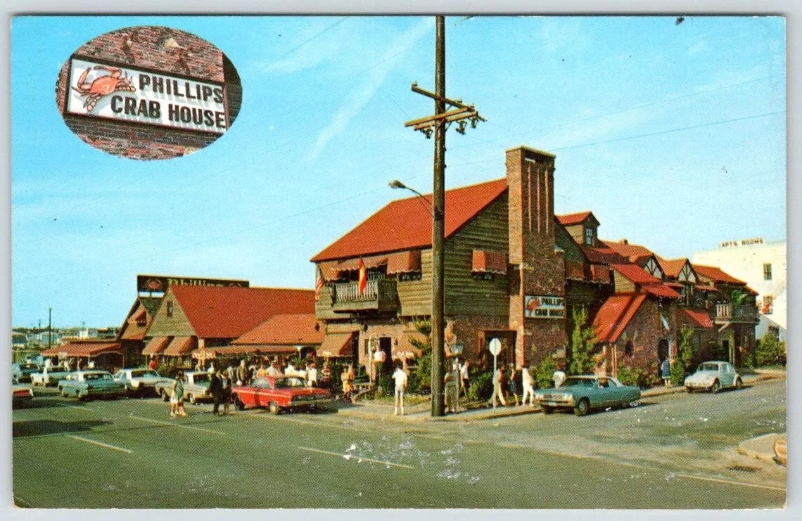  Vintage postcard of the Phillips Crab House exterior, a multi-building restaurant complex with red roofs and Tudor-style architecture, with cars and pedestrians visible in the parking area and a large oval Phillips Crab House sign prominently displa