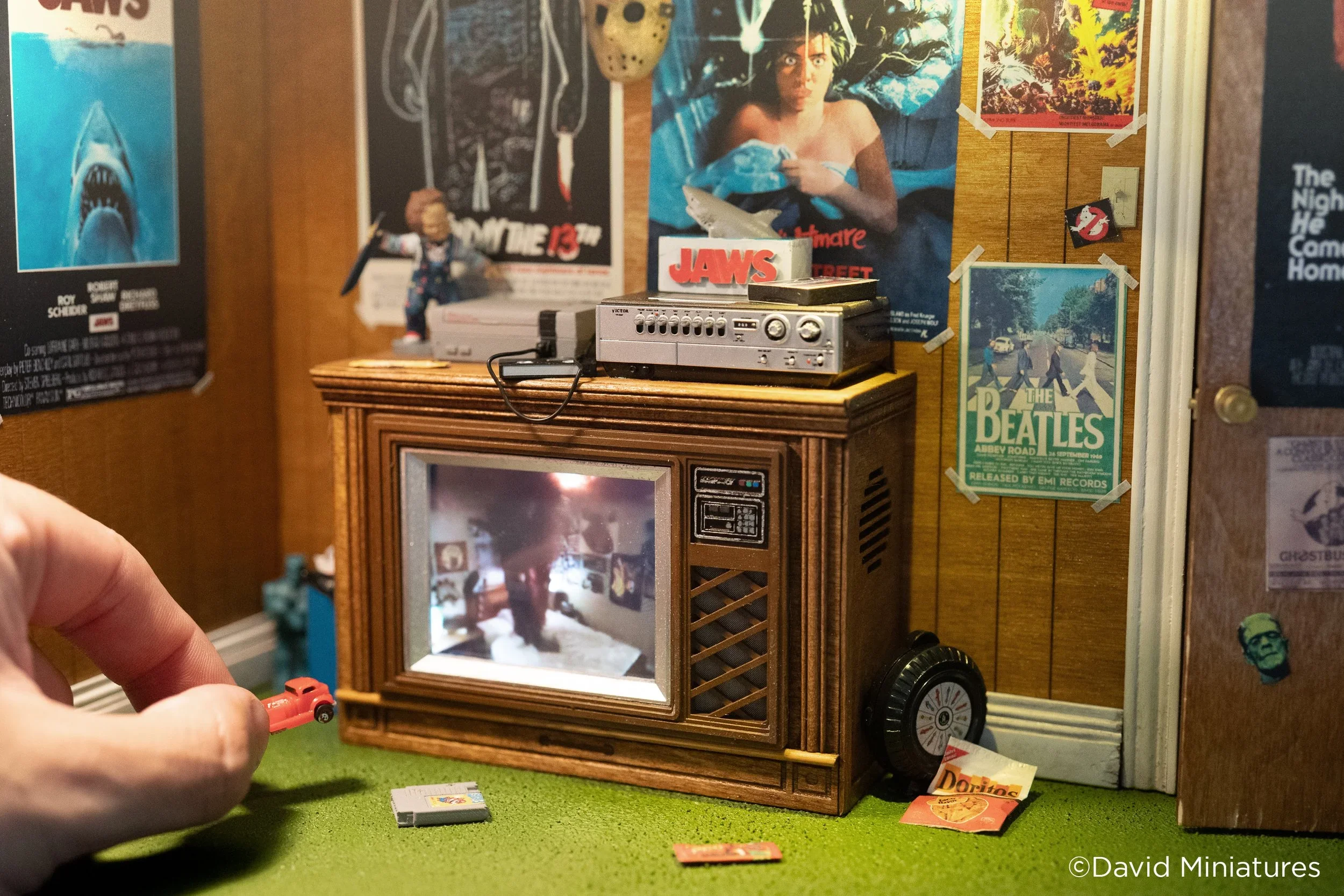 Miniature room with vintage TV, posters of movies and bands on the wall, a person holding a small red toy car, and various other tiny objects.
