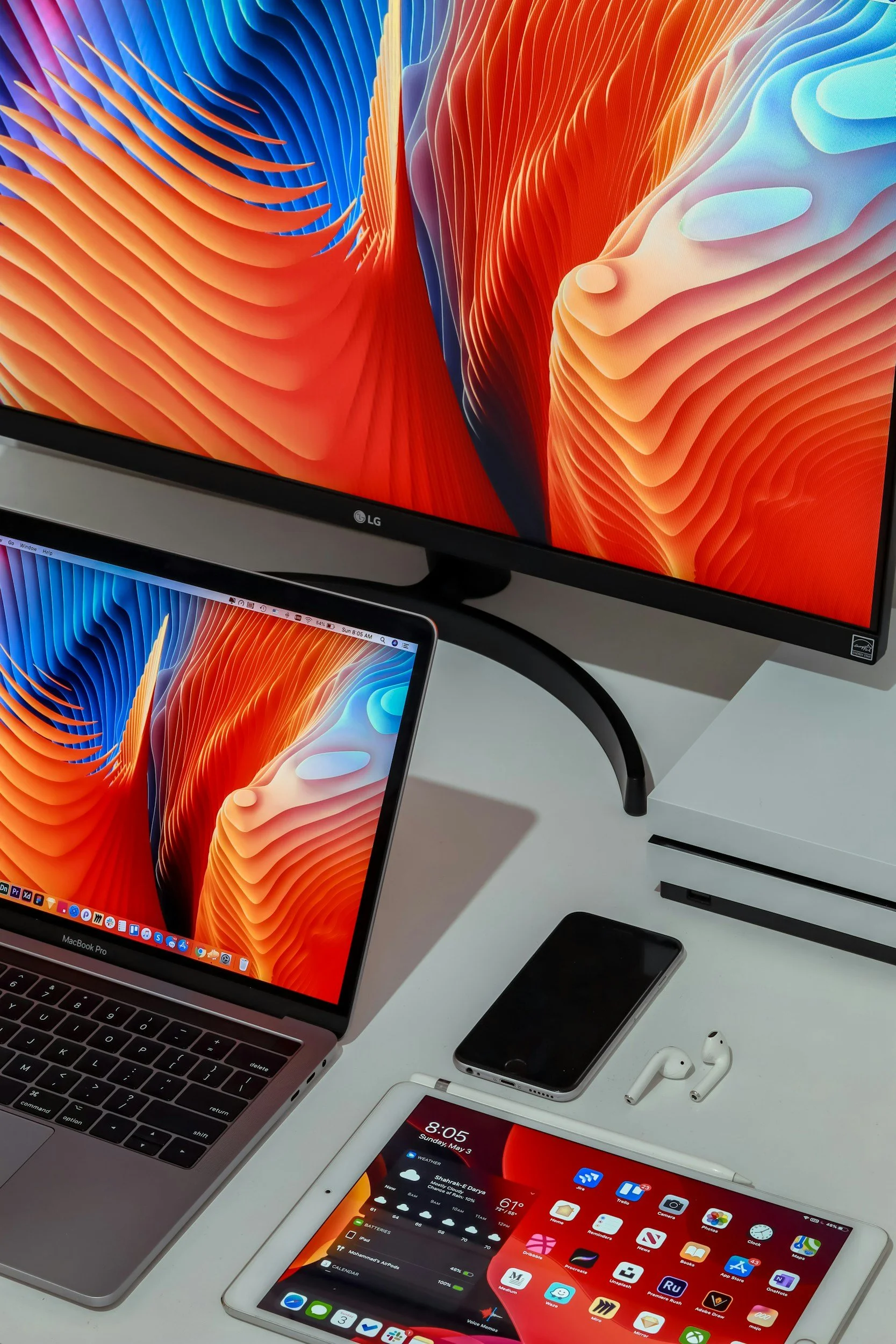 A workspace with Apple MacBook Pro laptop, iPad, iPhone, and Apple AirPods on a white desk, with a second monitor displaying a vibrant abstract wallpaper.