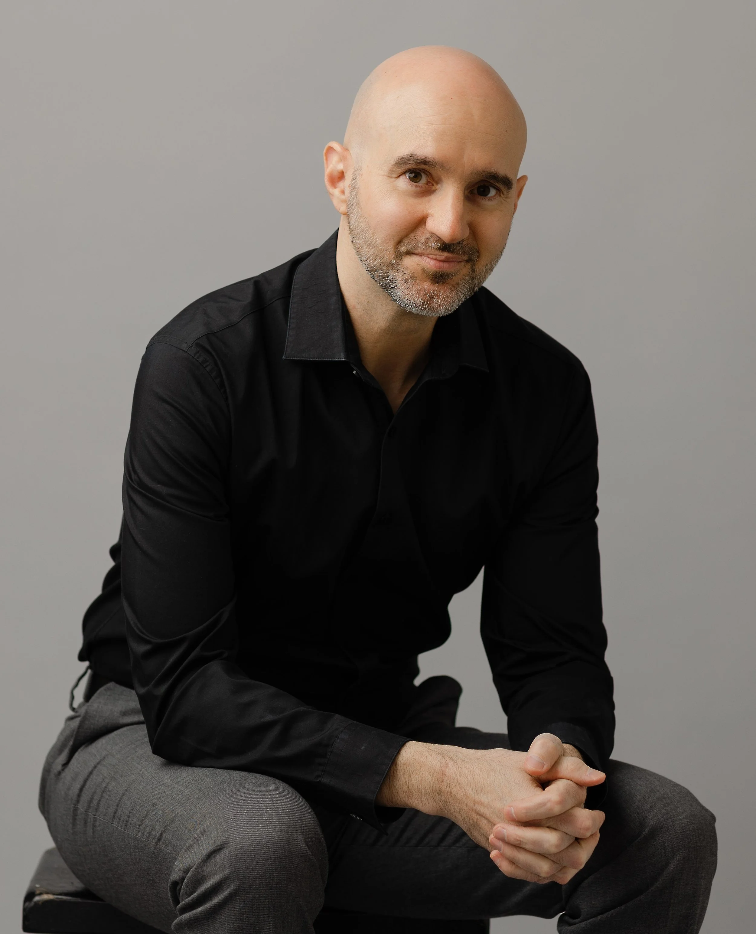 A bald man with a beard, wearing a black shirt and gray pants, sitting on a stool against a gray background, looking at the camera with his hands clasped.