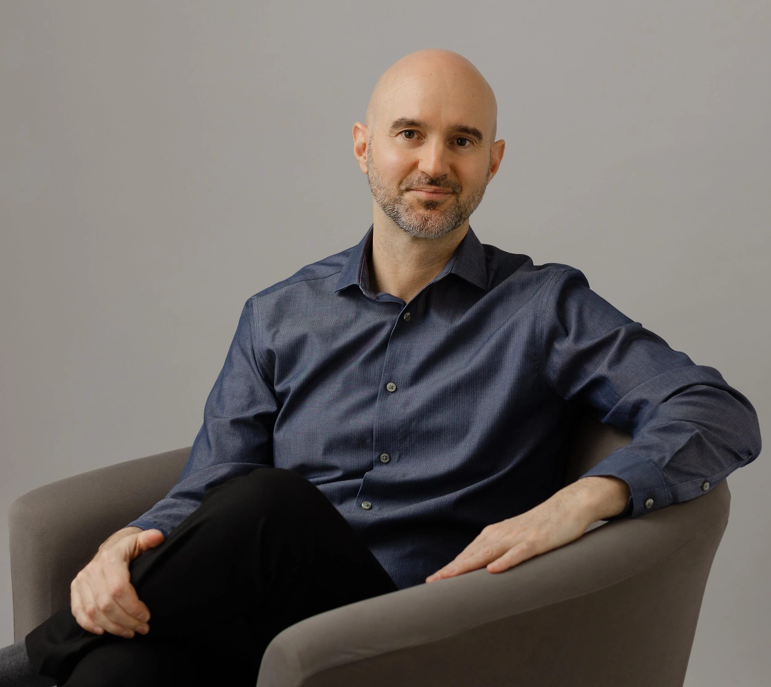A bald man with a beard wearing a dark blue button-up shirt sitting in a light gray armchair against a plain gray wall.
