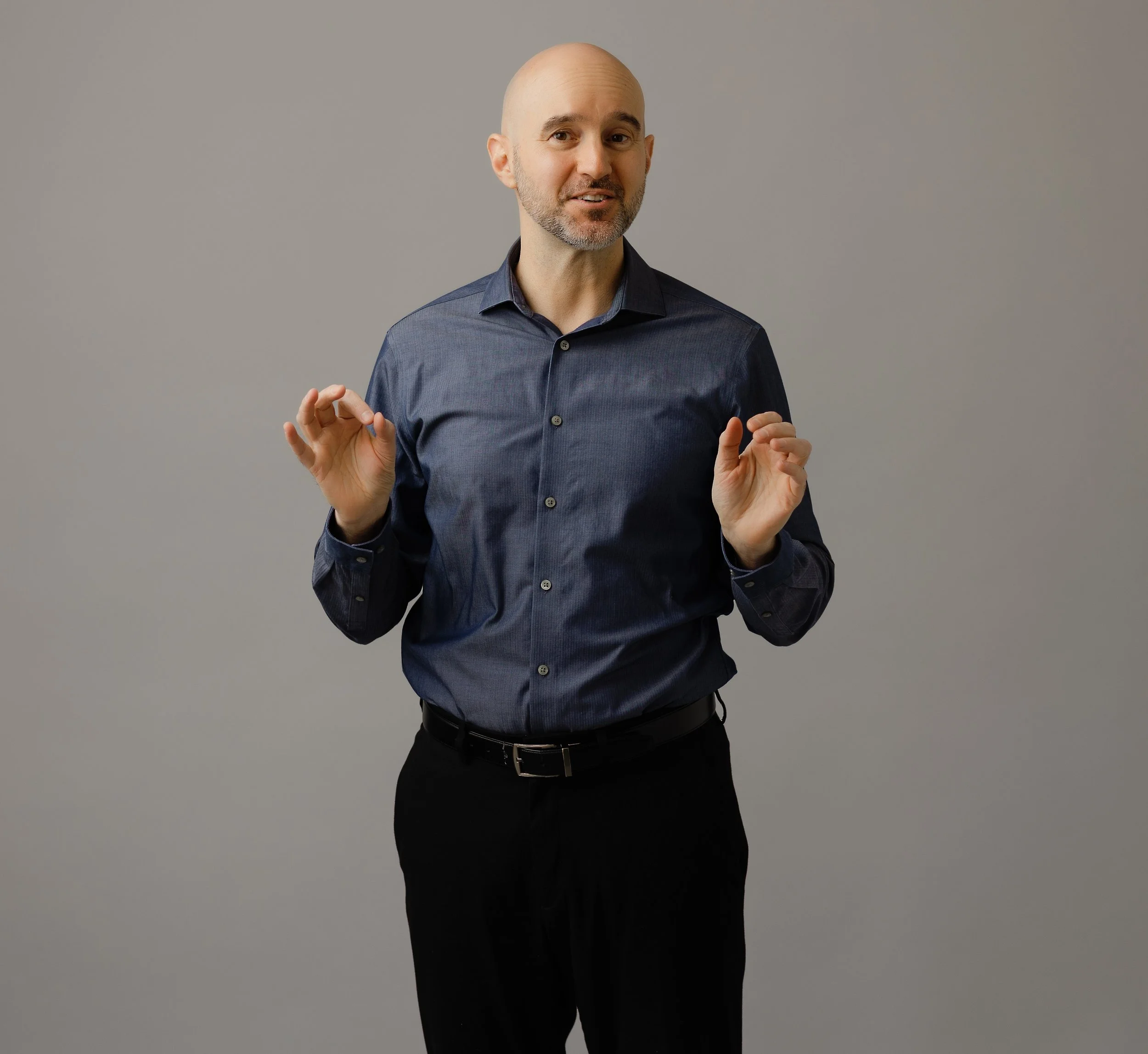 A man with a bald head and beard wearing a dark blue button-up shirt and black pants, standing against a gray background, making a gesture with both hands.