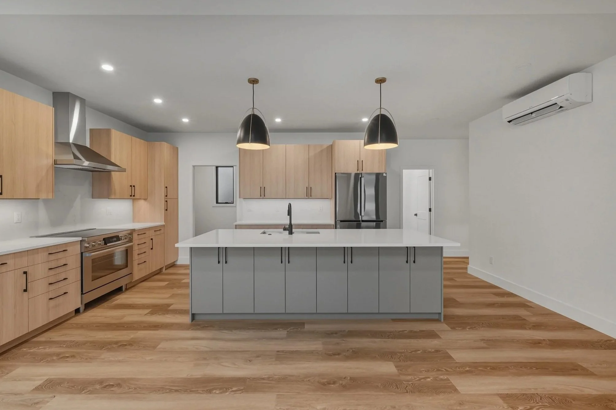 Modern empty kitchen with wooden cabinets, stainless steel appliances, and a central island with a white countertop, accompanied by pendant lights and hardwood flooring.