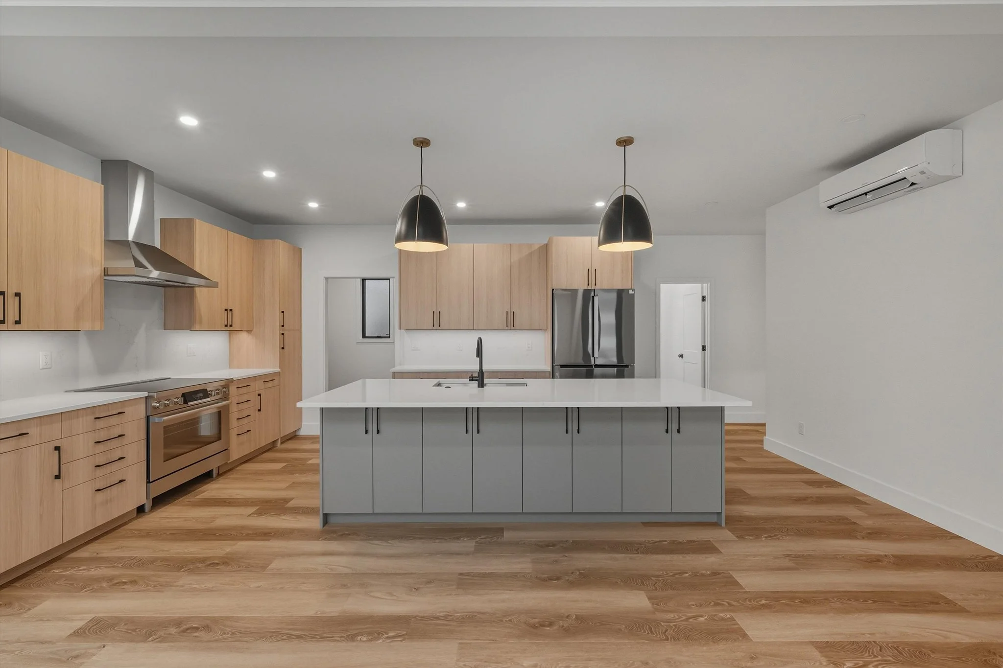Modern empty kitchen with wooden cabinets, stainless steel appliances, and a central island with a white countertop, accompanied by pendant lights and hardwood flooring.
