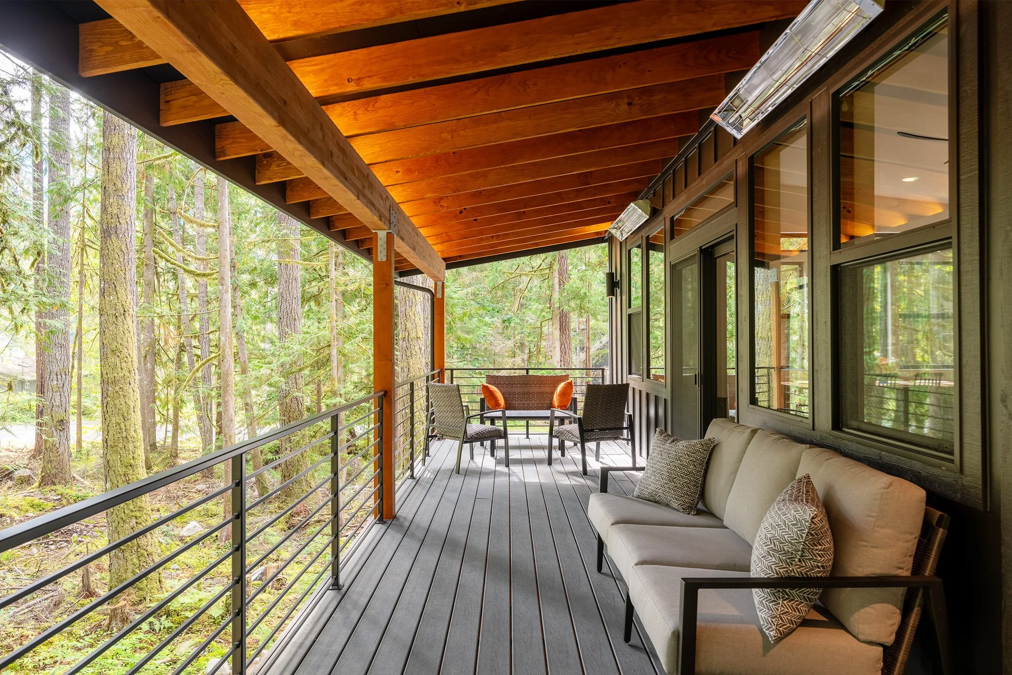 Covered outdoor balcony with a wooden ceiling, a cushioned outdoor sofa with patterned pillows, a table with four chairs, and a view of trees in the forest.
