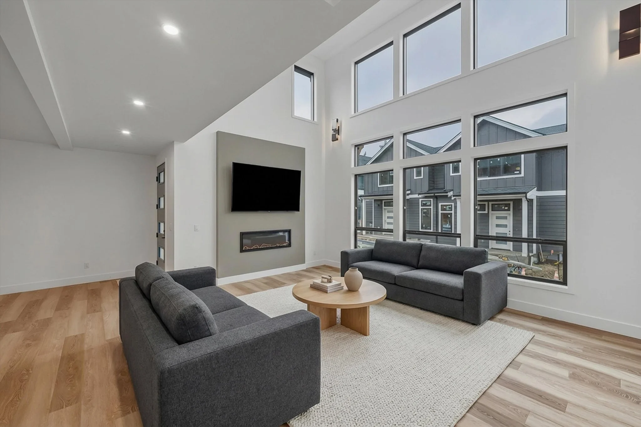 Modern living room with two gray sofas, a round wooden coffee table with books and a vase, large windows, a mounted TV, and a wall-mounted fireplace.