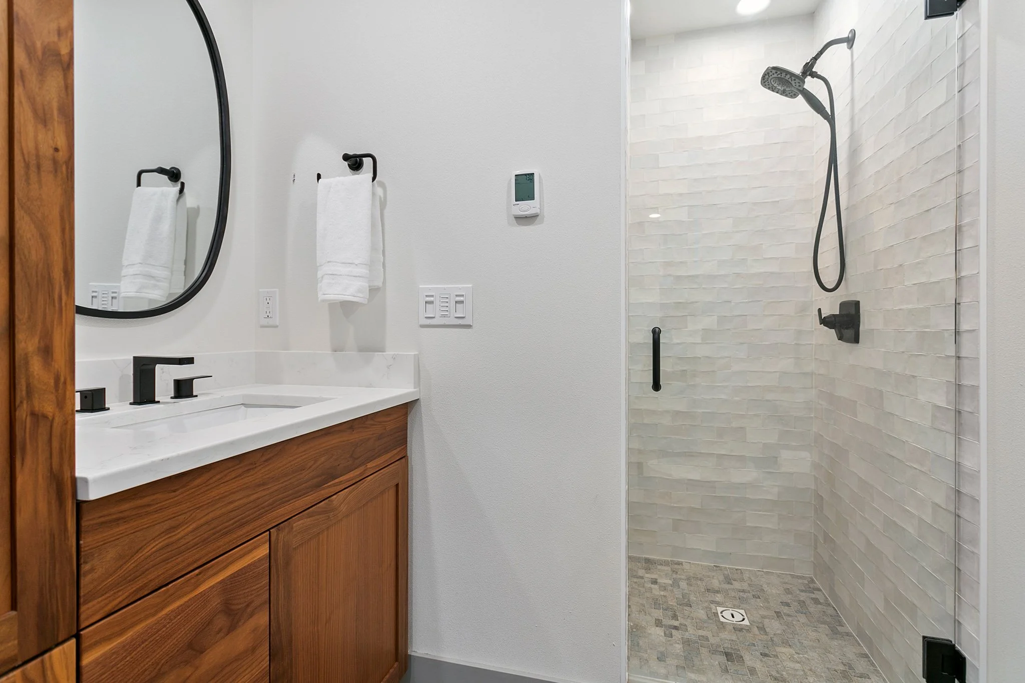 Modern bathroom with a wooden vanity, a white marble countertop, black fixtures, a large mirror, towel hooks with white towels, and a glass-enclosed shower with beige tile walls and a rainfall showerhead.