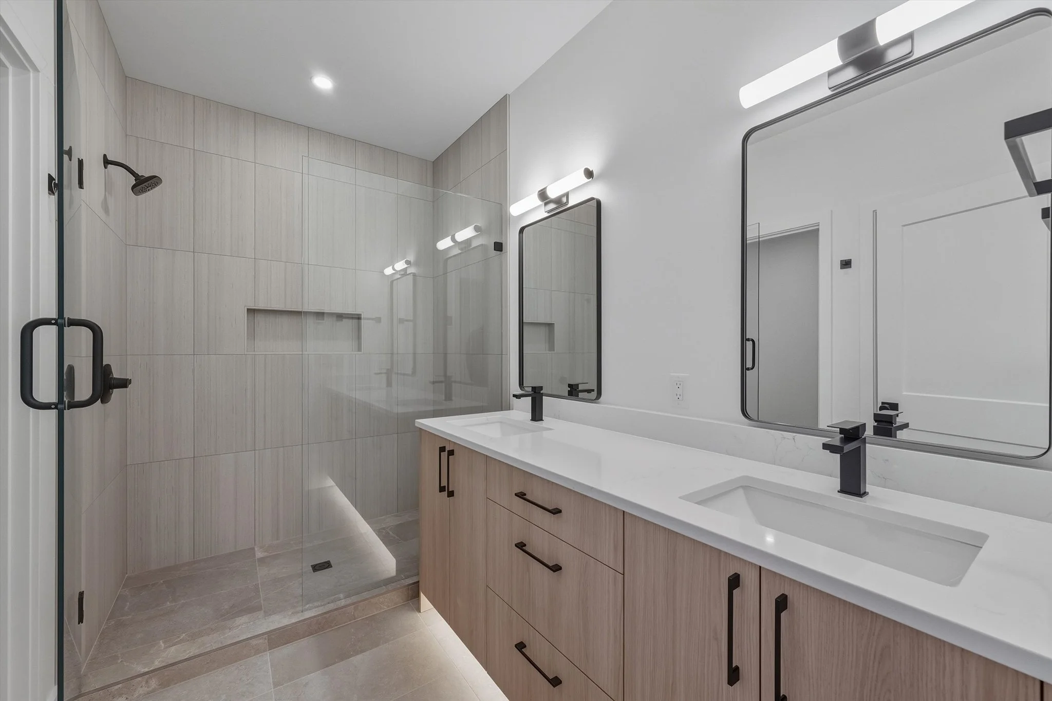 Modern bathroom with a double vanity sink, large mirrors, black fixtures, and a walk-in shower with a glass door and black handle, featuring beige tiled walls and floor.