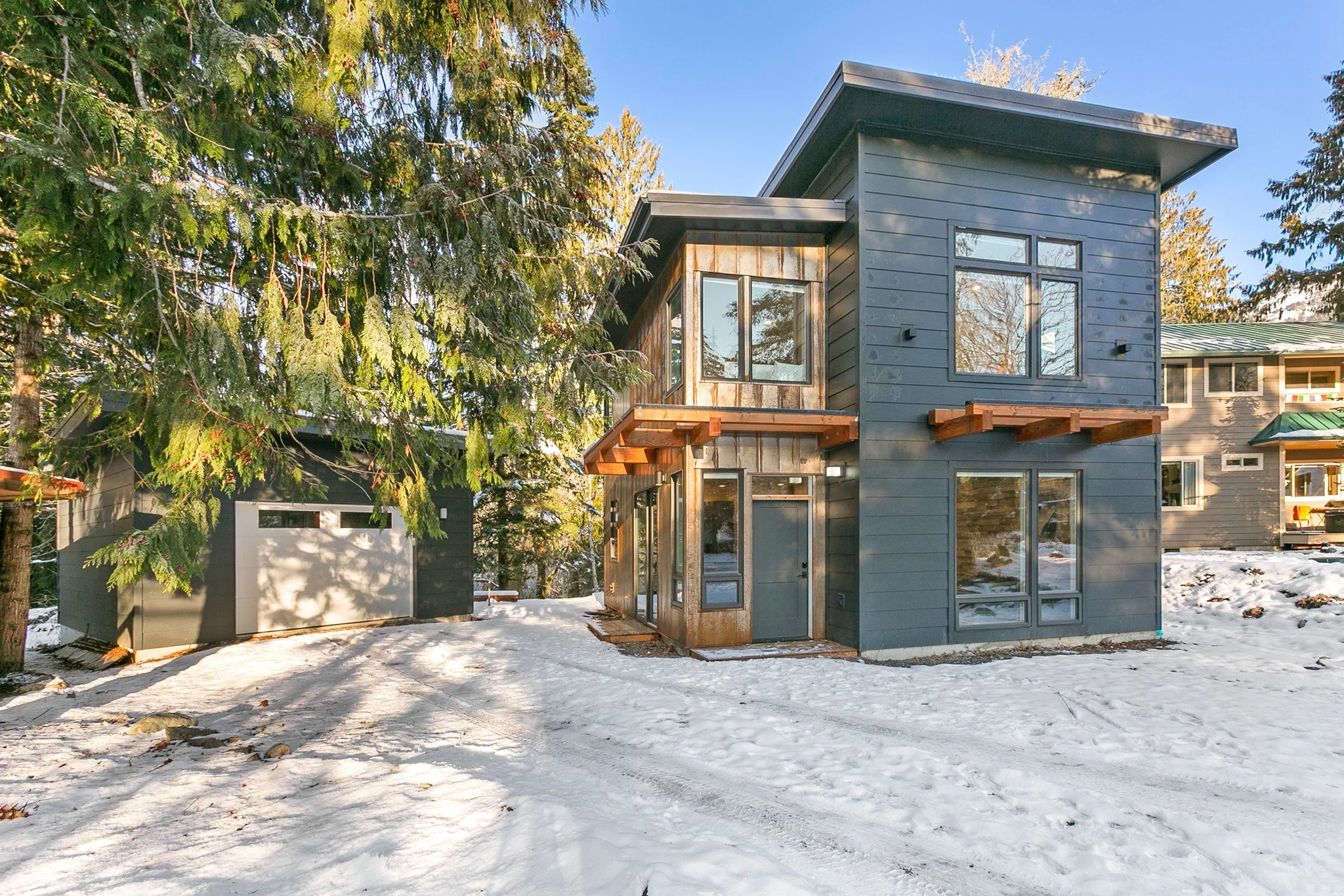 Modern house with a dark gray exterior and large windows, surrounded by snow and tall trees.