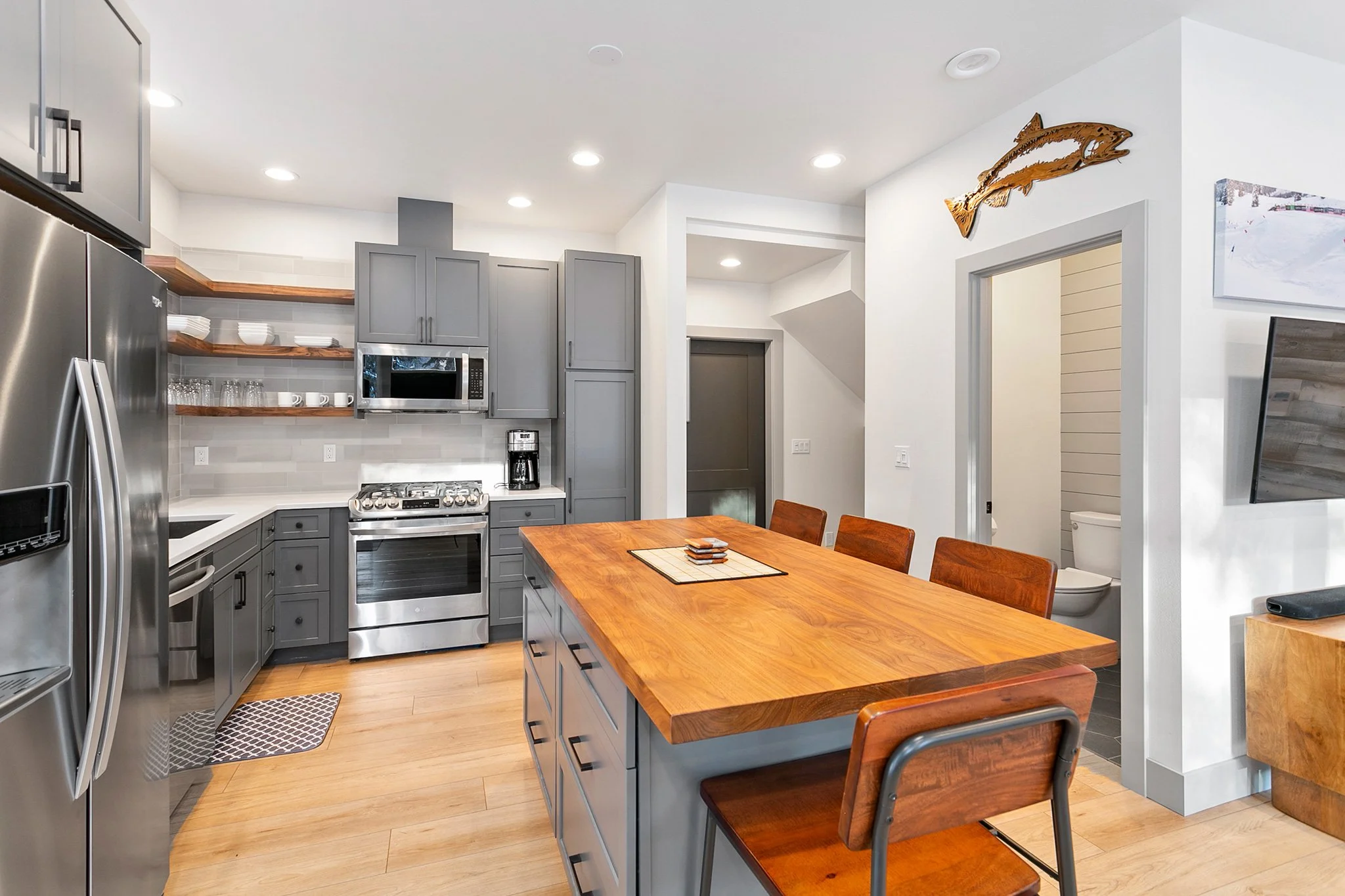 Modern kitchen with gray cabinets, wooden open shelves holding dishes and glassware, stainless steel appliances, a wooden dining table with matching chairs, and a small bathroom visible through an open door.