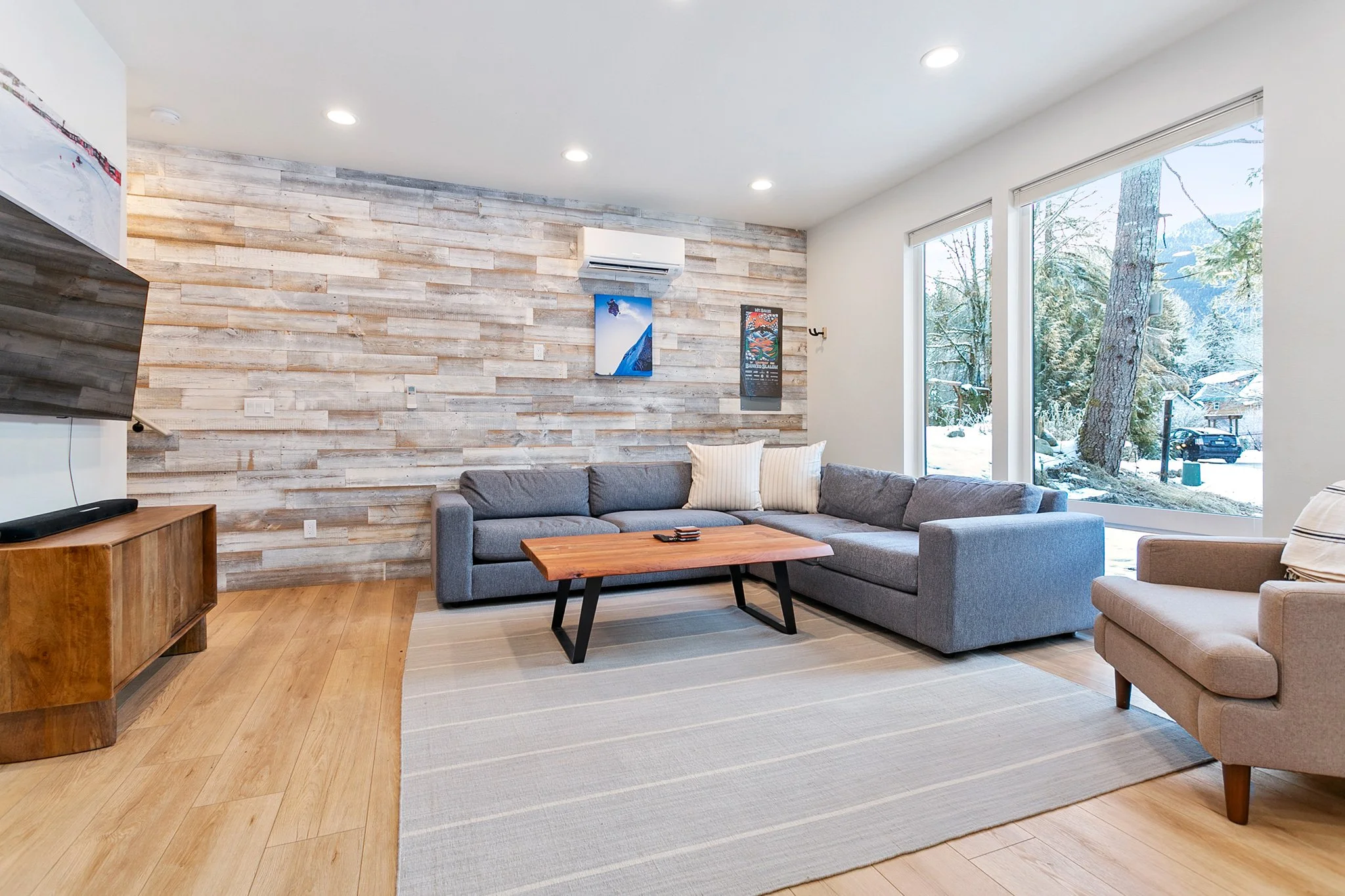 Living room with gray sectional sofa, wooden coffee table, beige armchair, wooden TV stand, wall art, large windows showing snowy outdoor scene, wood flooring, recessed ceiling lights.