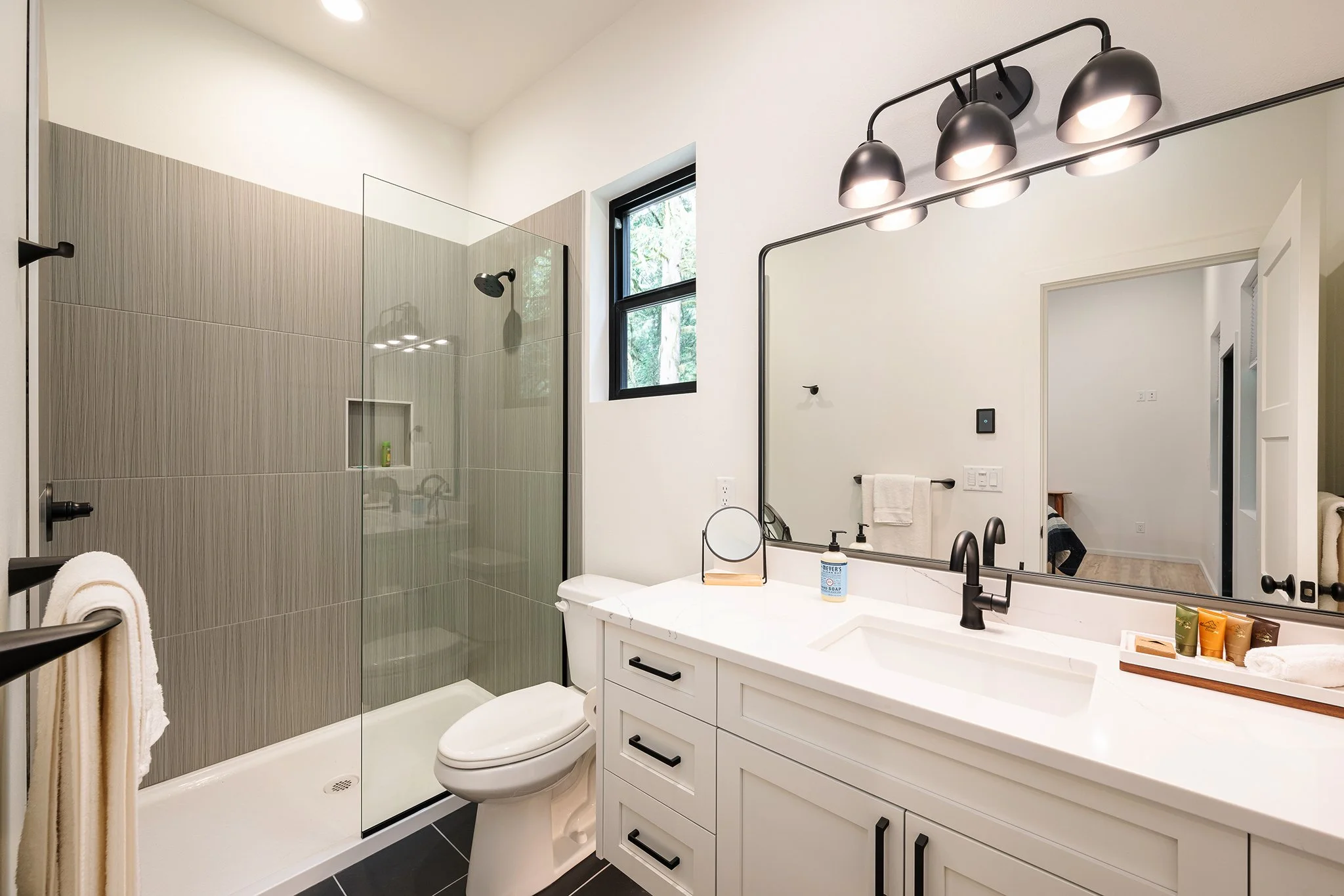 Modern bathroom with walk-in shower, white vanity with black hardware, large mirror, black light fixture, and window with trees outside