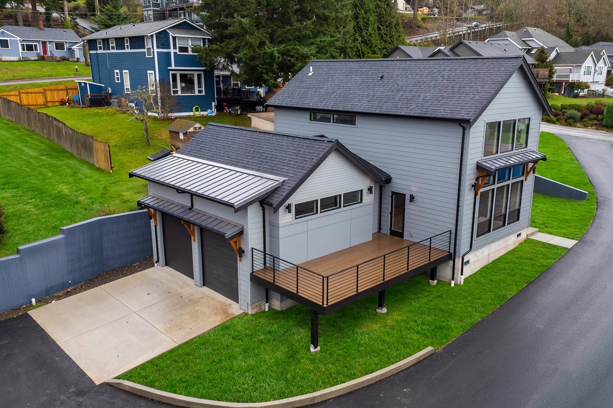 A modern two-story house with a gray exterior, blue siding, and a metal roof, featuring a wooden deck, a garage with black doors, and surrounded by green lawns and neighboring houses.