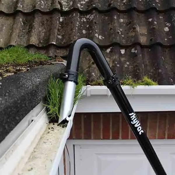 A leaf blower in black and silver color being used to blow leaves and dirt off a gutter on the edge of a roof.