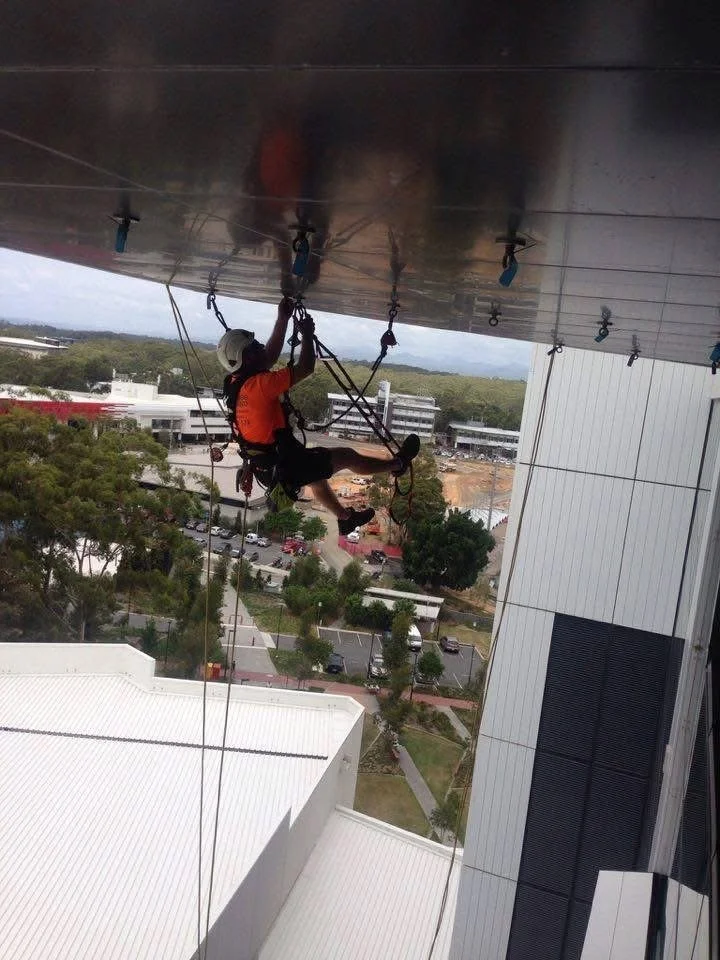 A window cleaner is suspended high up on the exterior of a tall building, wearing safety gear, to clean the glass window.